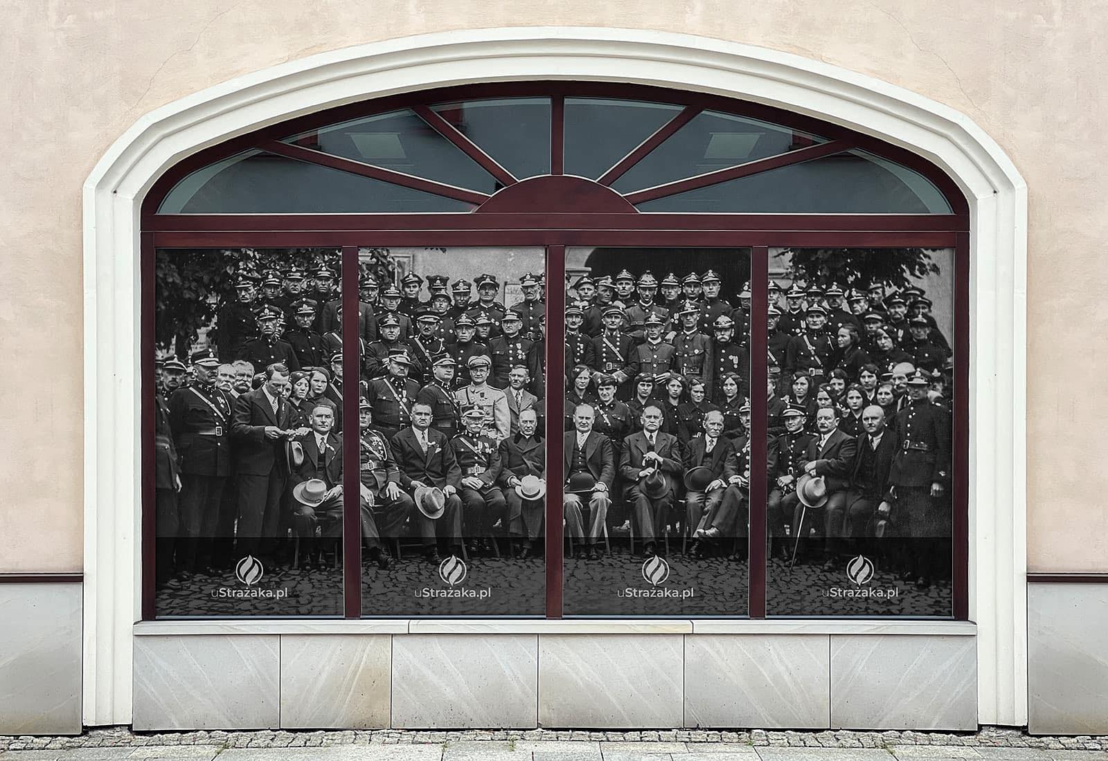 Czarno-biała fotografia dużej grupy umundurowanych strażaków i osób cywilnych umieszczona na okiennej folii reklamowej z adresem uStrazaka.pl.