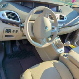 Jasne, beżowe wnętrze samochodu Renault Scenic z widoczną deską rozdzielczą, kierownicą z logo, fotelem kierowcy i dźwignią zmiany biegów.