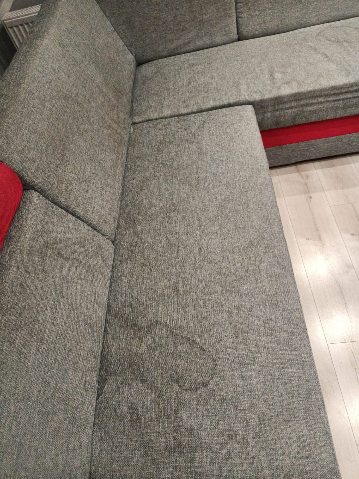 Szara, narożna kanapa z widocznym, okrągłym zabrudzeniem na siedzisku, z czerwonym akcentem na dole i jasną podłogą w tle.