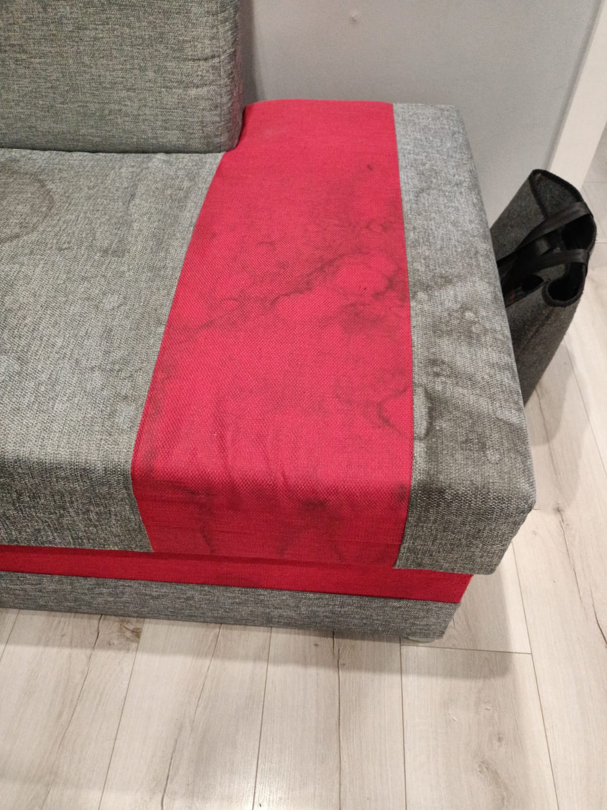Fragment szarej kanapy z czerwoną wstawką, widoczne plamy na czerwonym materiale, obok stoi szara torba na zakupy.