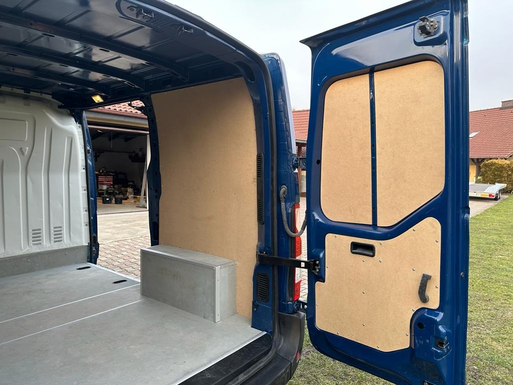 Wnętrze niebieskiego furgonetki wyłożone płytami MDF, z otwartymi tylnymi drzwiami i widocznym garażem w tle.