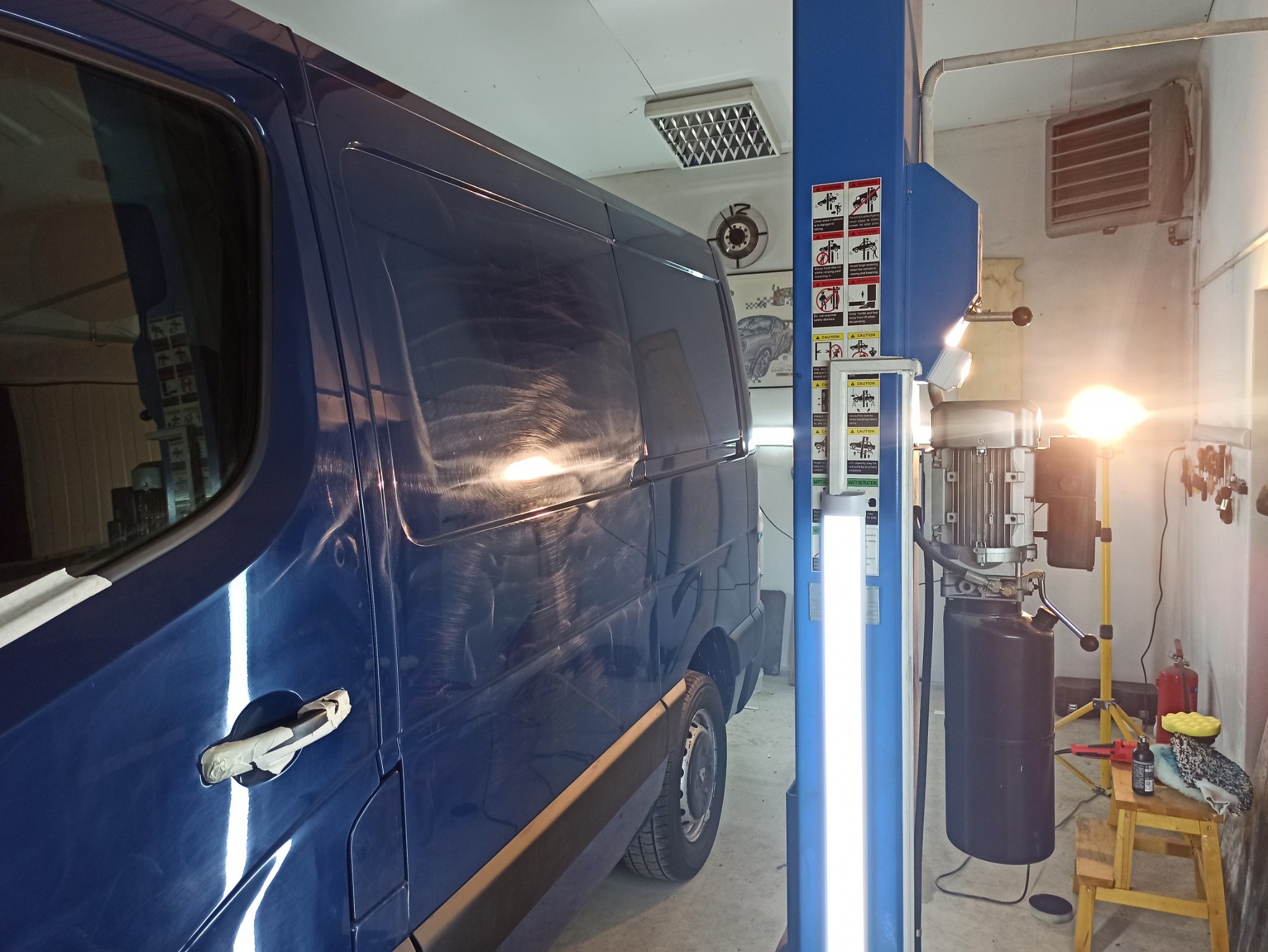 Niebieski van częściowo owinięty taśmą malarską, stojący w warsztacie samochodowym obok podnośnika, przygotowywany do polerowania lakieru.