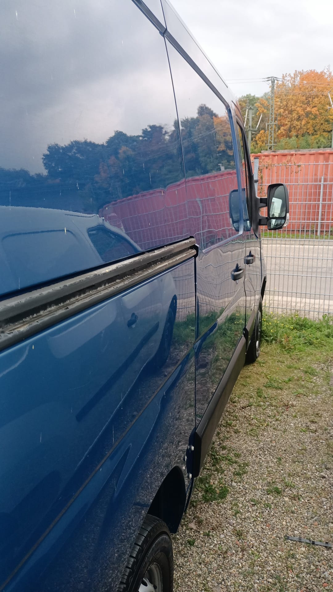 Boczny widok niebieskiego samochodu dostawczego z widocznymi kroplami deszczu na karoserii, zaparkowanego na żwirowym podłożu, z odblaskiem drzew i nieba na lakierze.