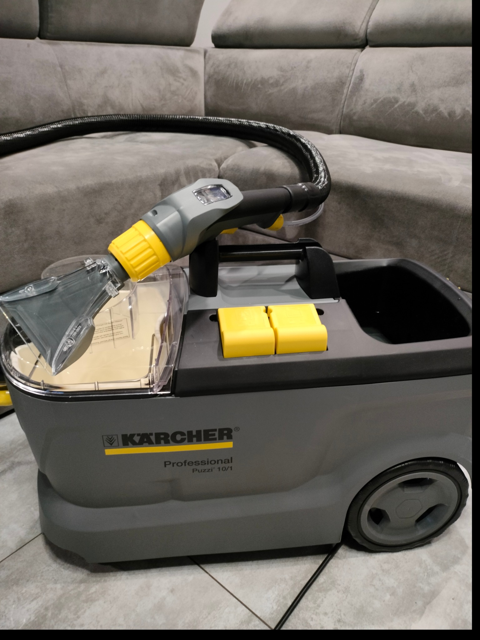 Profesjonalny odkurzacz piorący Kärcher Puzzi 10/1 z przezroczystym zbiornikiem na detergent, dyszą i wężem, ustawiony na tle szarej kanapy.