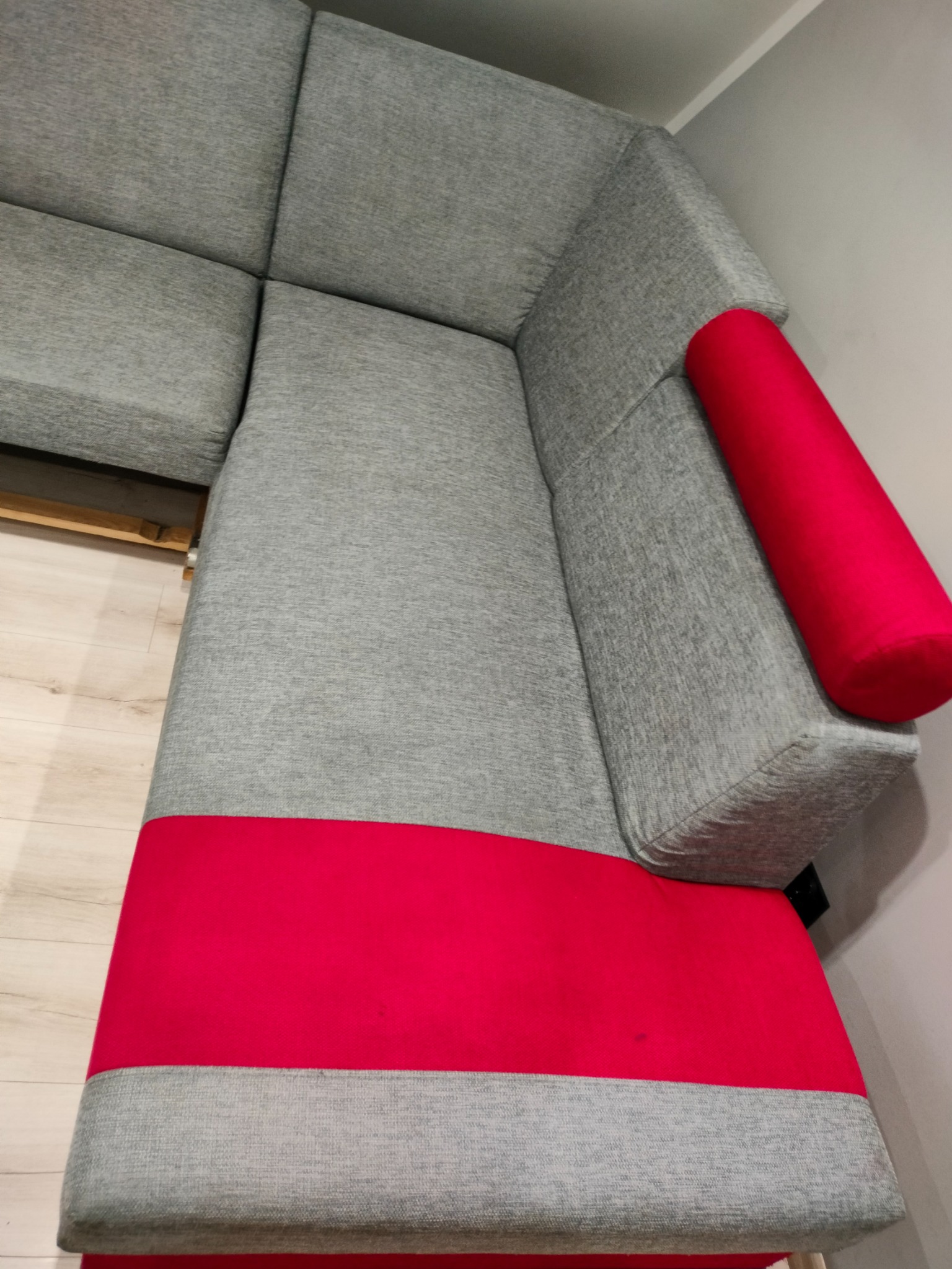 Szary narożnik z czerwoną wstawką na siedzisku i czerwoną poduszką wałkiem, widok z góry pod kątem.
