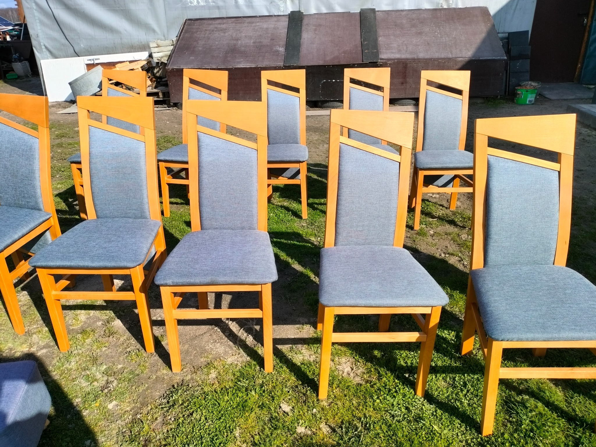 Rząd krzeseł z drewnianymi ramami i szarą tapicerką, ustawionych na trawie przed budynkiem gospodarczym.