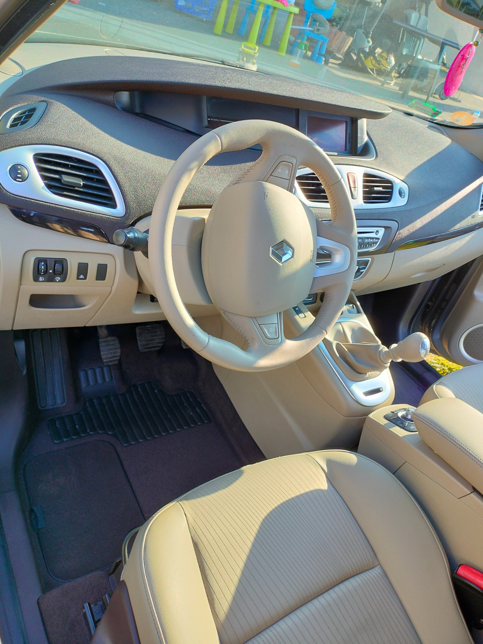 Jasne, beżowe wnętrze samochodu Renault Scenic z widoczną deską rozdzielczą, kierownicą z logo, fotelem kierowcy i dźwignią zmiany biegów.