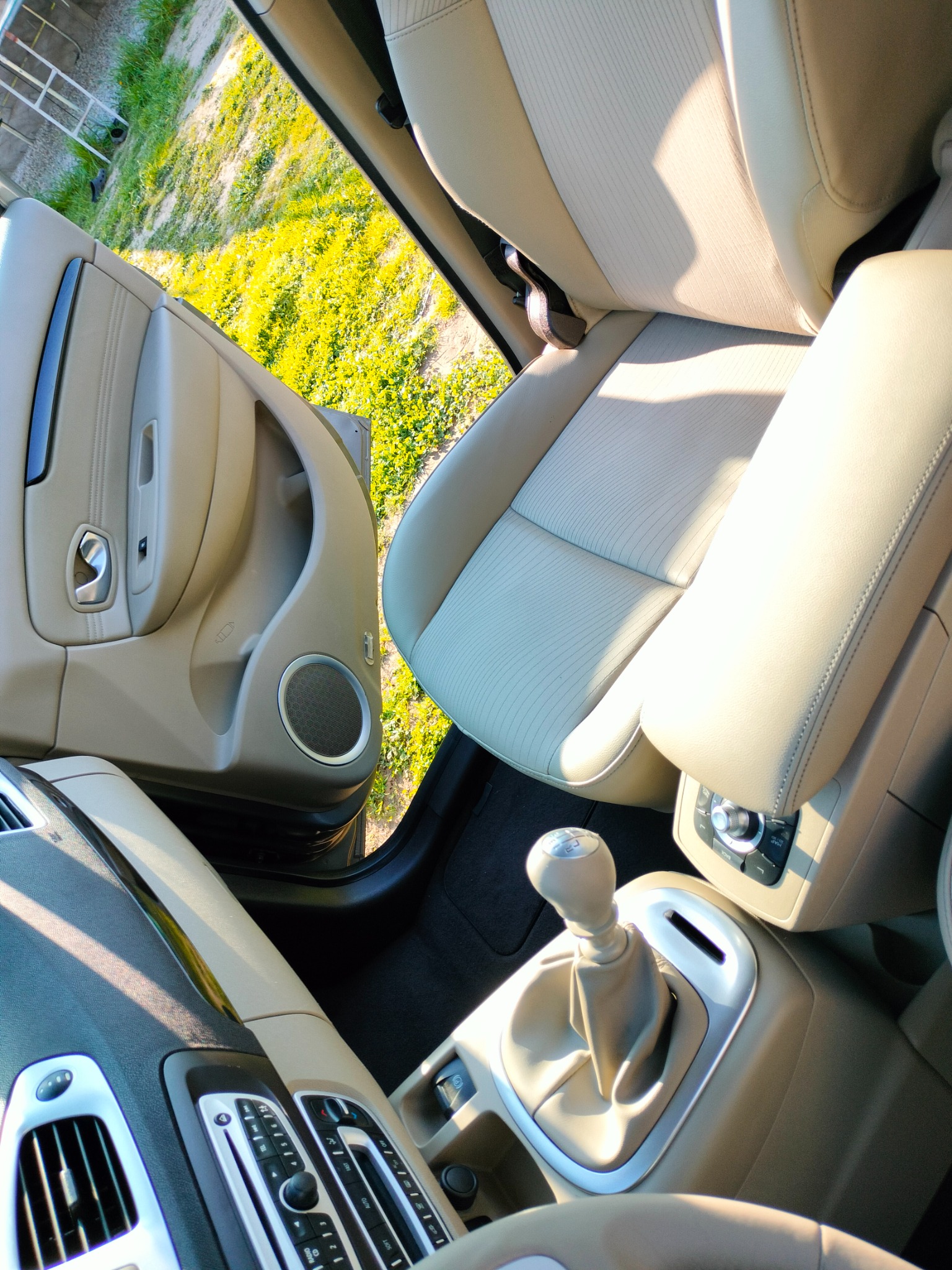 Wnętrze samochodu z beżową tapicerką, widoczne fotele, konsola środkowa z dźwignią zmiany biegów, panel sterowania i otwarte drzwi z widokiem na zieleń.