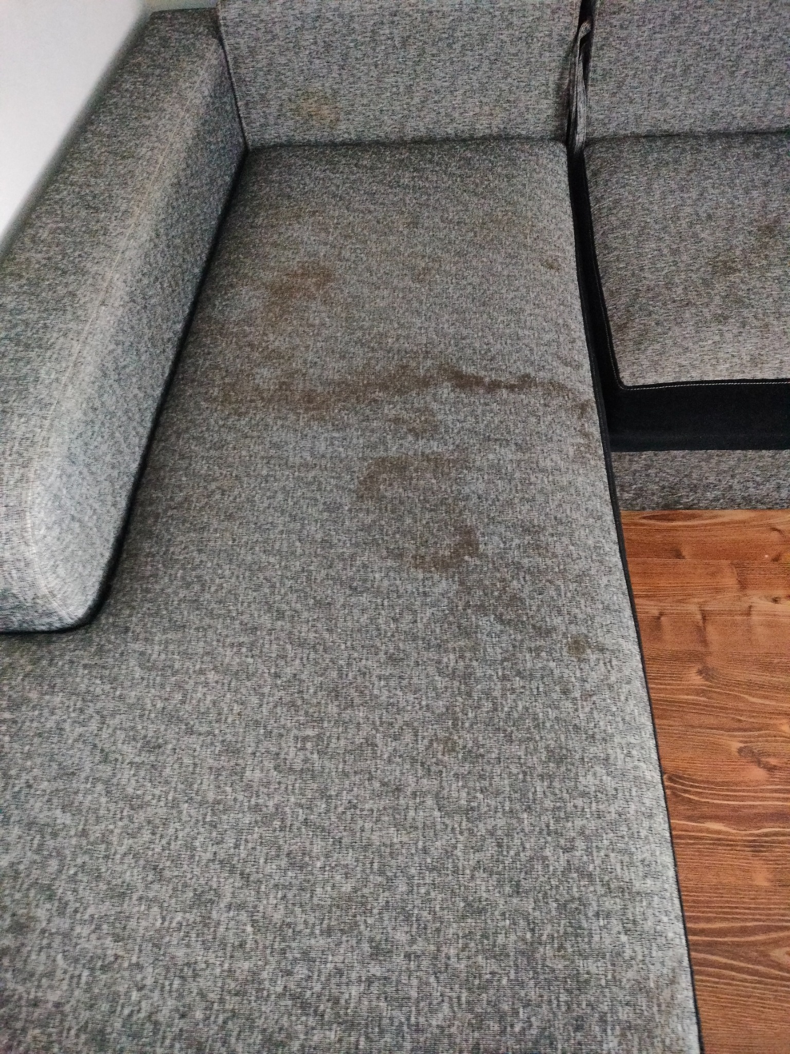 Szara, narożna kanapa z widocznymi plamami na siedzisku i oparciu, stojąca na drewnianej podłodze.