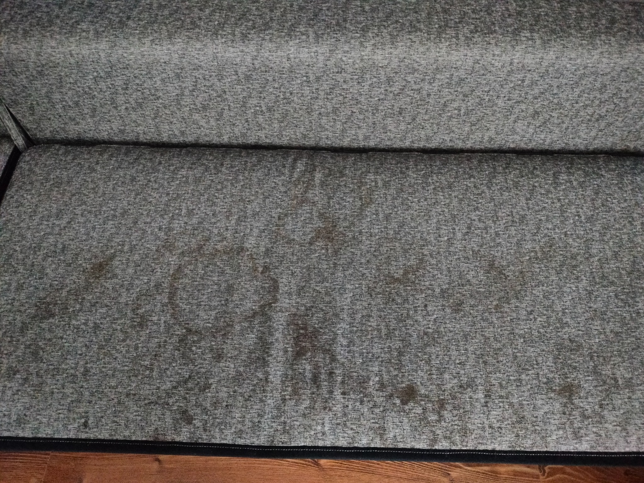 Fragment szarej kanapy z widocznymi plamami i zabrudzeniami na siedzisku, z drewnianą podłogą w tle.