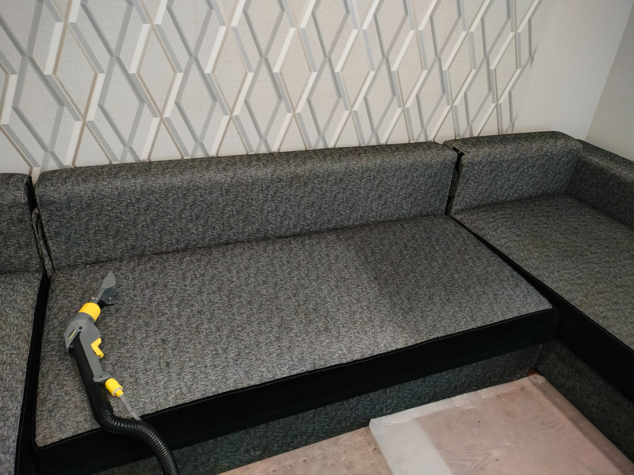Szara kanapa narożna z czarnymi obiciami czyszczona odkurzaczem piorącym z żółtymi elementami, wzorzysta tapeta w tle.
