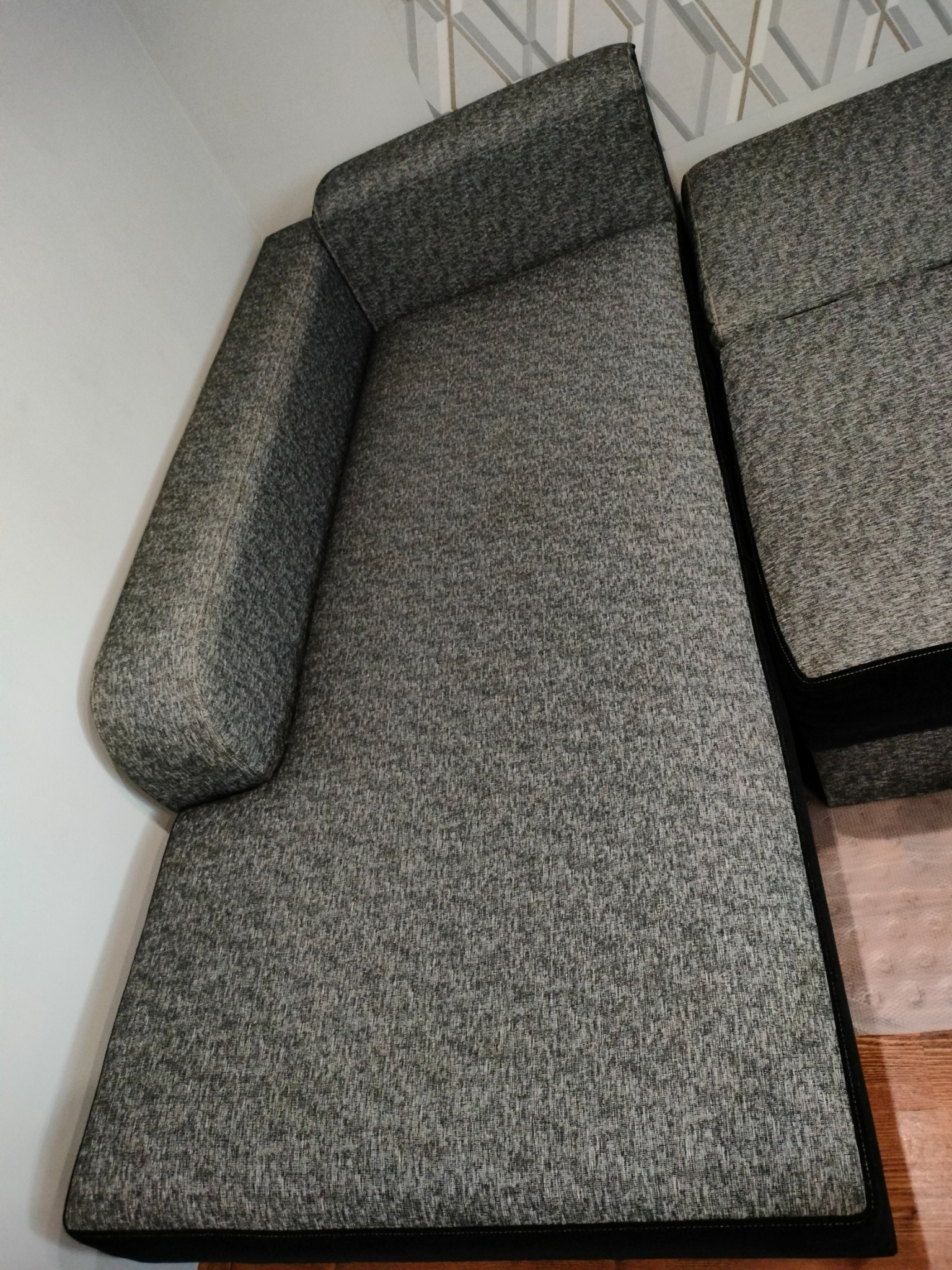 Szara kanapa narożna z tapicerką o teksturze melanżowej, widoczna z góry, z poduszką boczną i czarnym obramowaniem podstawy.