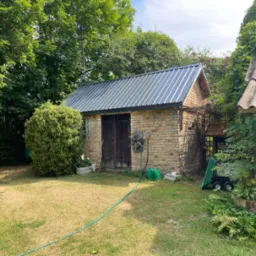 Murowana szopa gospodarcza z blachy falistej na dachu, otoczona zielenią i z wężem ogrodowym na trawniku.