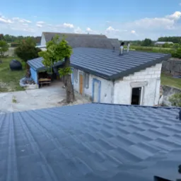 Widok z góry na nowo położony, ciemnoszary dach z blachy trapezowej na budynku gospodarczym z białego bloczka, z widocznymi kominami i obróbkami blacharskimi, w tle inne budynki i błękitne niebo...