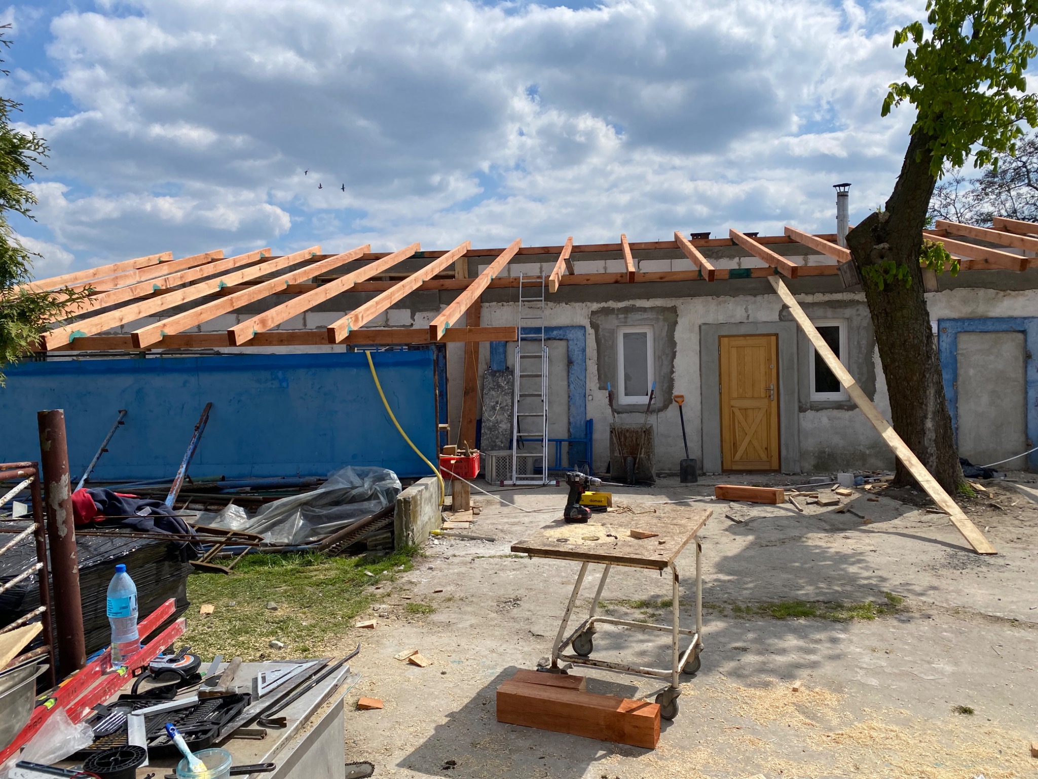 Wznoszenie więźby dachowej z drewnianych belek na murowanym budynku z niebieskim akcentem, widoczne narzędzia i drabina w tle, drzewo obok budynku.