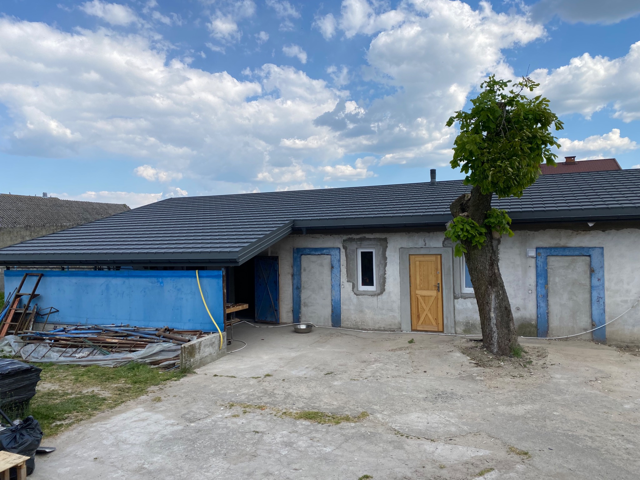 Nowy, grafitowy dach z blachodachówki na parterowym budynku gospodarczym, widoczne nieotynkowane ściany, drewniane drzwi i podwórze z nieporządkiem.