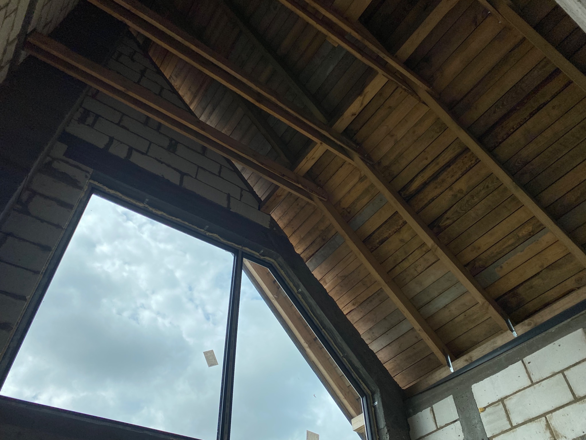 Wnętrze surowego domu w trakcie budowy, widoczna więźba dachowa z drewnianych belek, ściany z białych bloczków, okno z widokiem na pochmurne niebo.