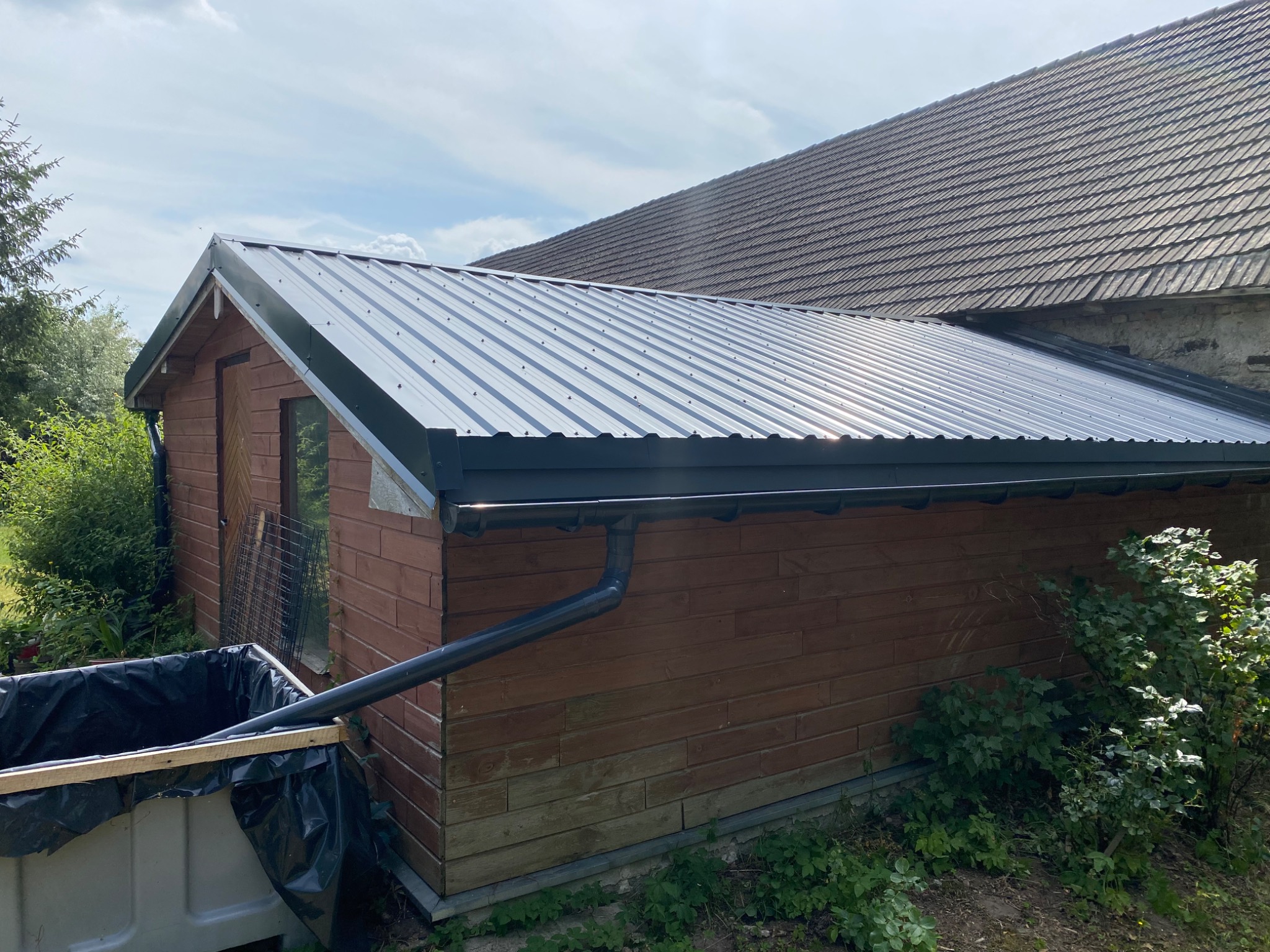 Nowy, blaszany dach z orynnowaniem na drewnianej przybudówce, z widocznym fragmentem starego domu z dachówką w tle.