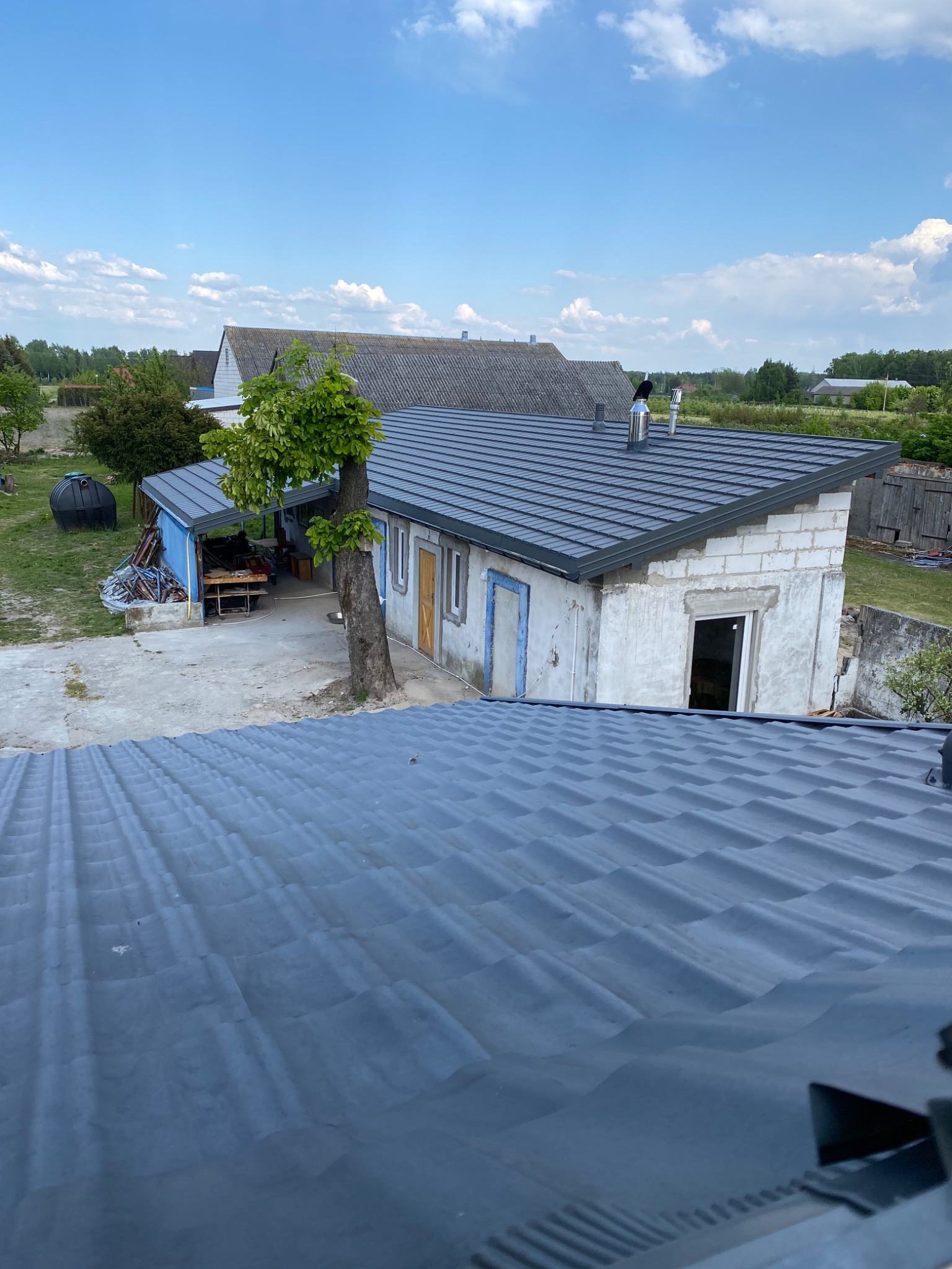 Widok z góry na nowo położony, ciemnoszary dach z blachy trapezowej na budynku gospodarczym z białego bloczka, z widocznymi kominami i obróbkami blacharskimi, w tle inne budynki i błękitne niebo...