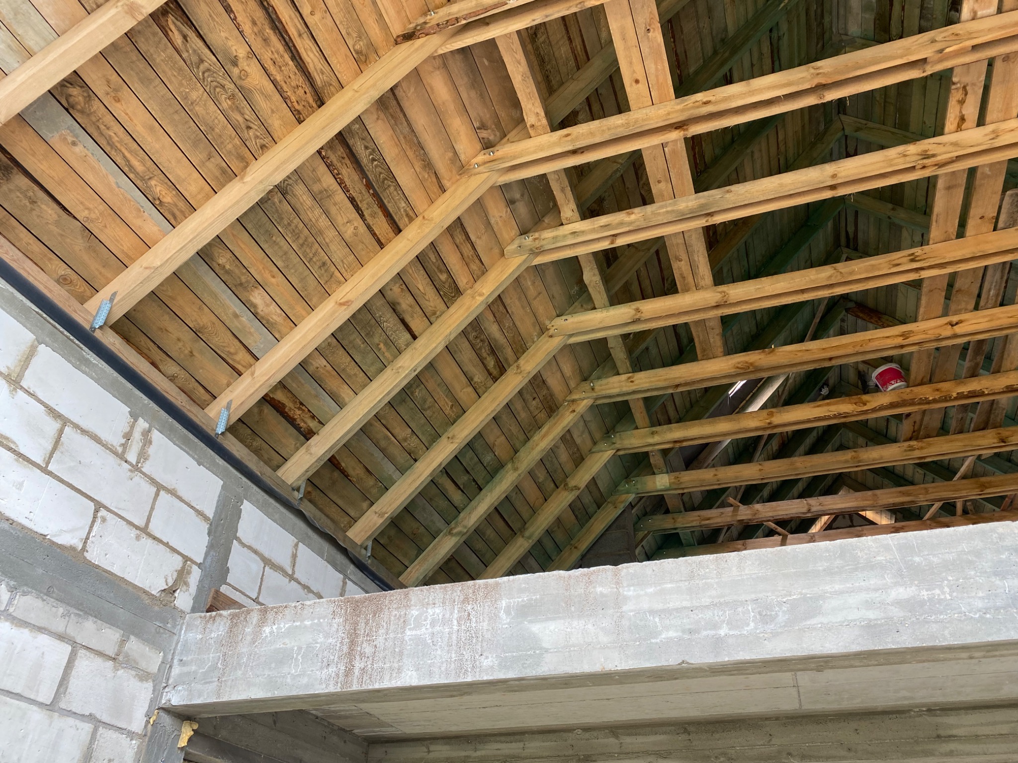 Wnętrze budynku w trakcie budowy, widoczna drewniana konstrukcja dachu z elementami mocującymi i fragment murowanej ściany z betonu komórkowego.