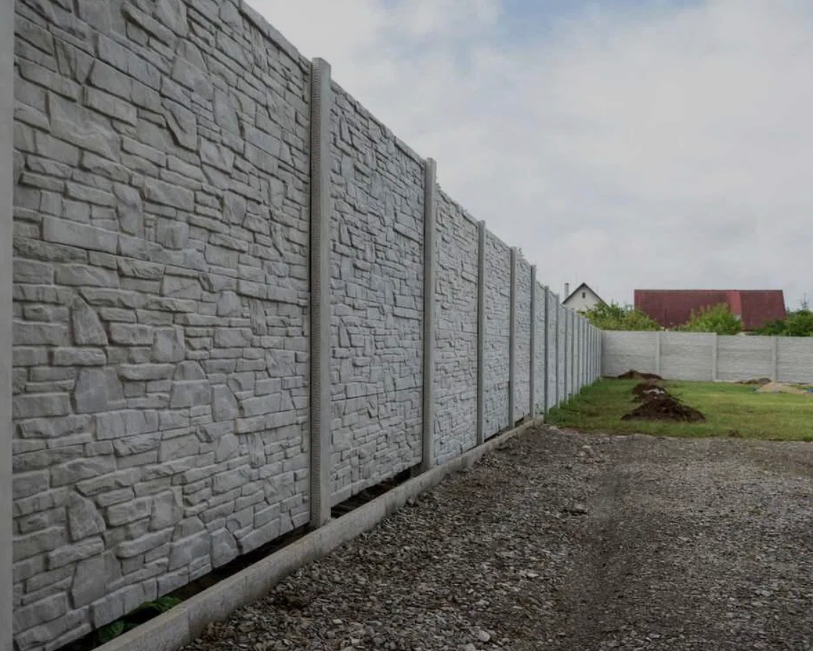 Długi, prefabrykowany betonowy płot z teksturą kamienia, z widocznymi słupkami i podmurówką, oddzielający posesję od drogi żwirowej, z domem w tle.