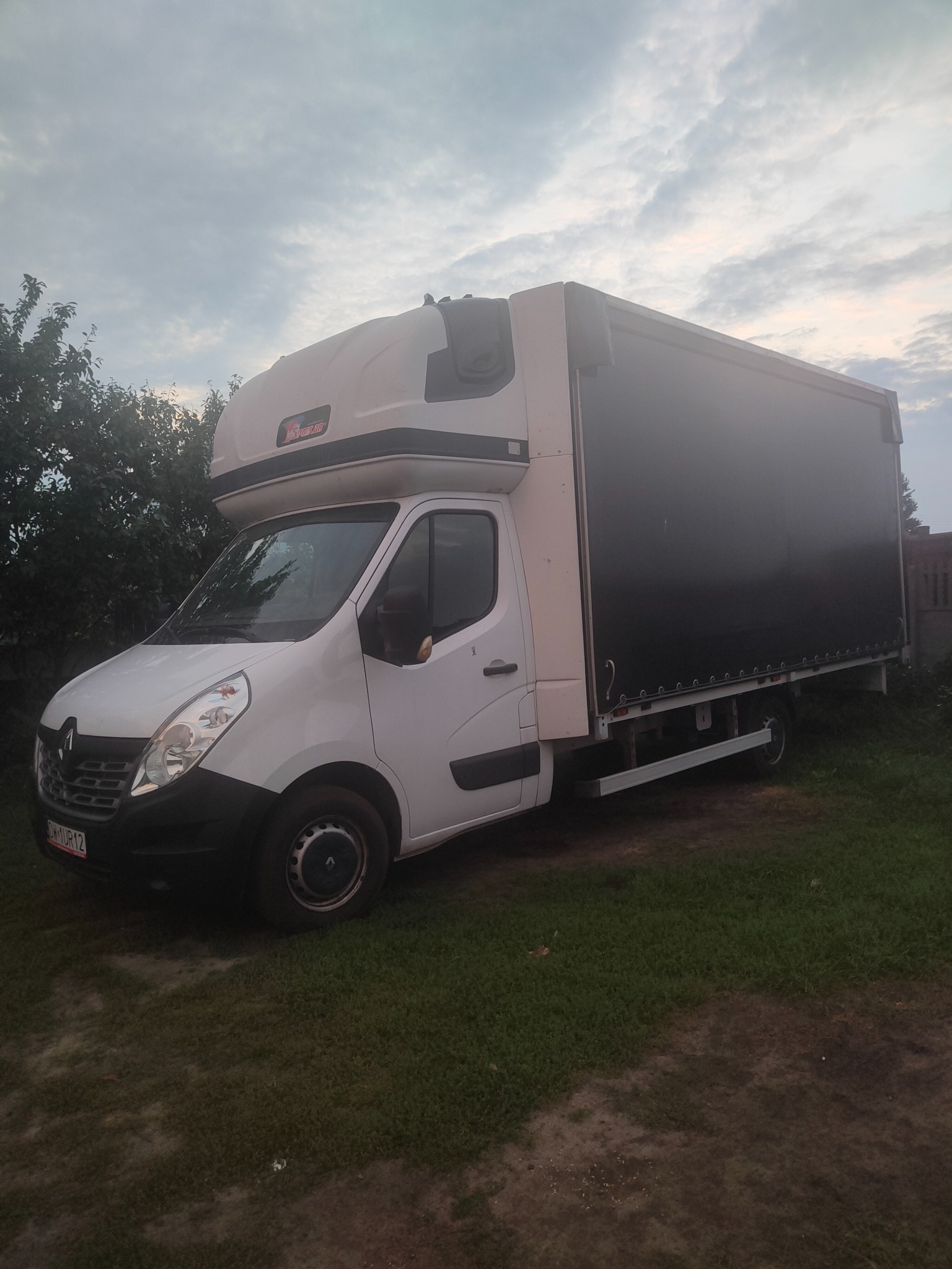 Biały samochód dostawczy Renault Master z plandeką na trawiastym podłożu, widok z boku, tablica rejestracyjna widoczna.