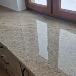 Polerowany blat z kamienia o beżowo-brązowej barwie, zamontowany przy oknie z drewnianą ramą, widok na zimowy krajobraz za oknem, fragment szafki z uchwytami widoczny w dolnej części zdjęcia.