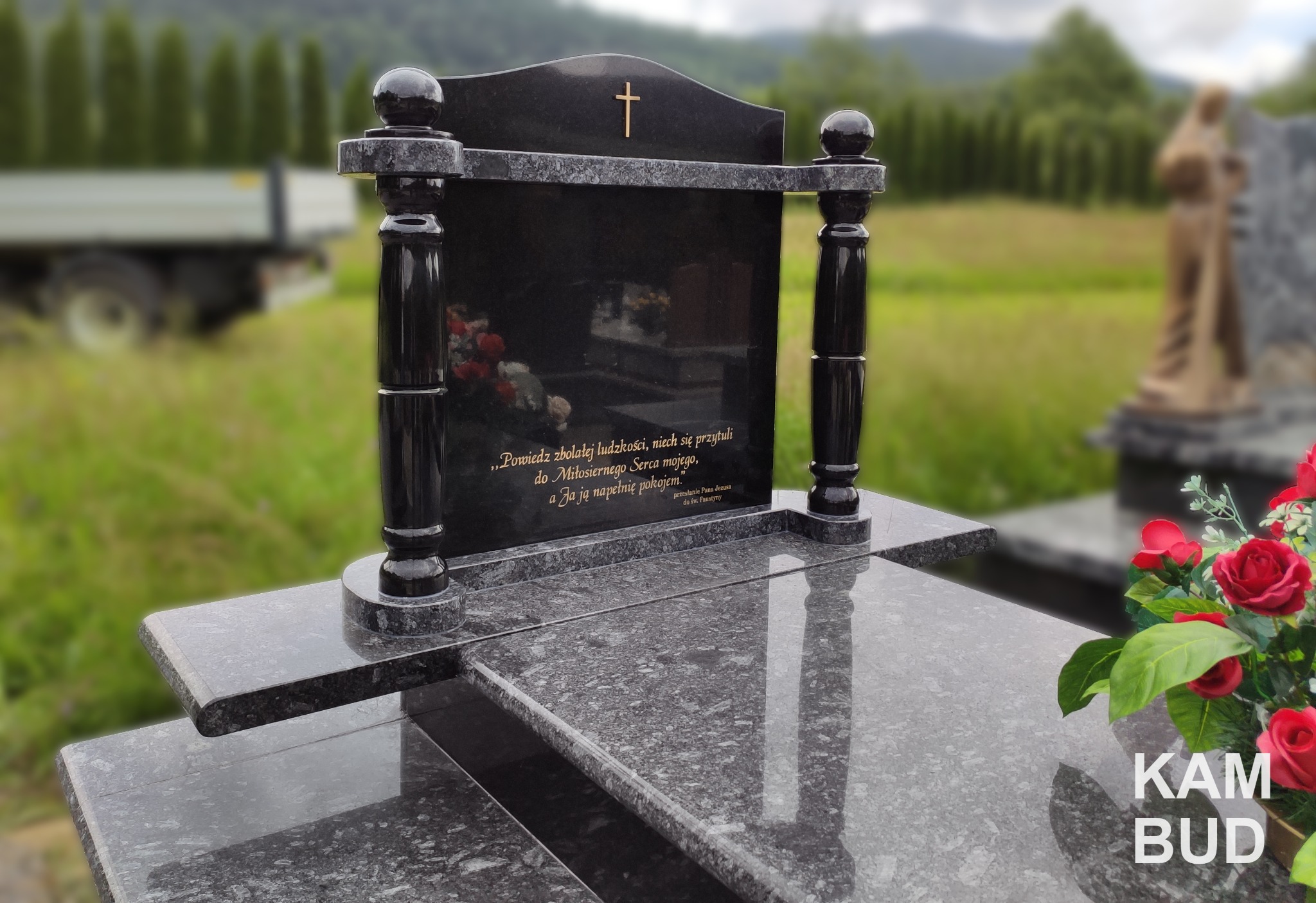 Czarny, polerowany nagrobek z granitu z inskrypcją, zwieńczony złotym krzyżem, na tle zieleni i rozmytego krajobrazu. Kompozycja uzupełniona sztucznymi czerwonymi różami.