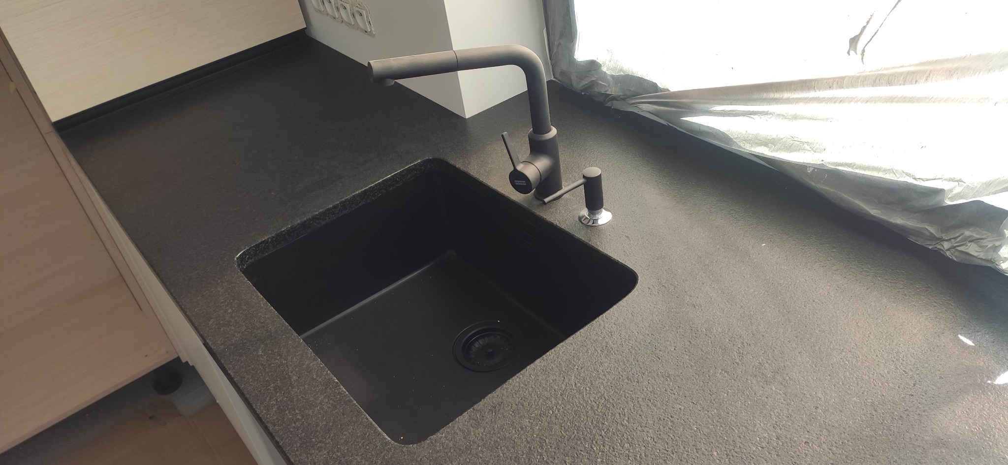 Czarny, matowy zlew kuchenny z baterią i dozownikiem na tle blatu o ciemnej, ziarnistej fakturze, częściowo osłoniętego folią ochronną.