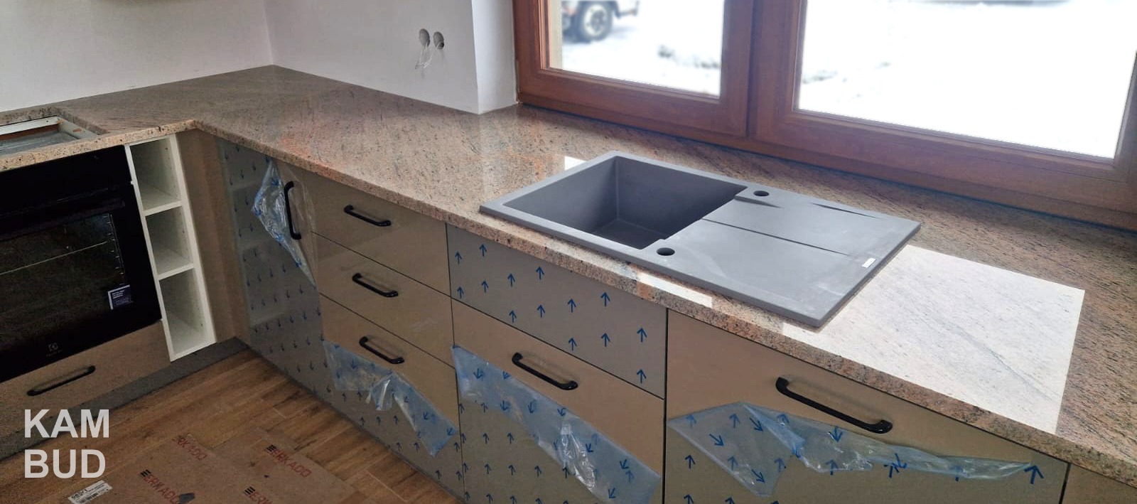 Beżowo-brązowy blat z kamienia z szarą, prostokątną umywalką, zamontowany w kuchni z folią ochronną na frontach szafek.