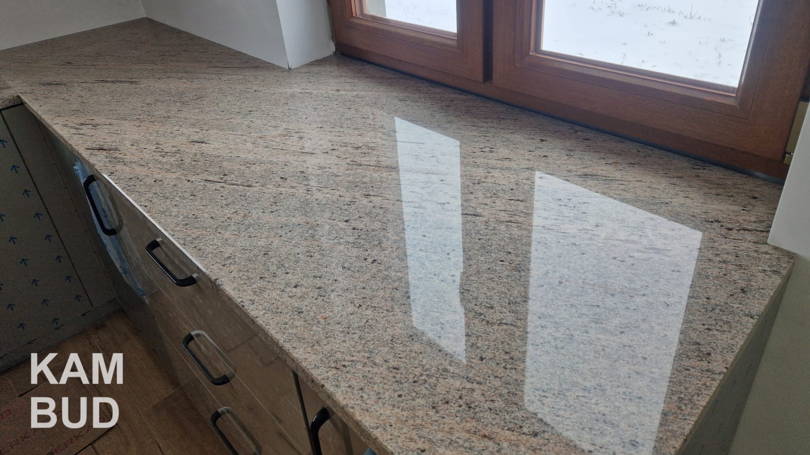 Polerowany blat z kamienia o beżowo-brązowej barwie, zamontowany przy oknie z drewnianą ramą, widok na zimowy krajobraz za oknem, fragment szafki z uchwytami widoczny w dolnej części zdjęcia.