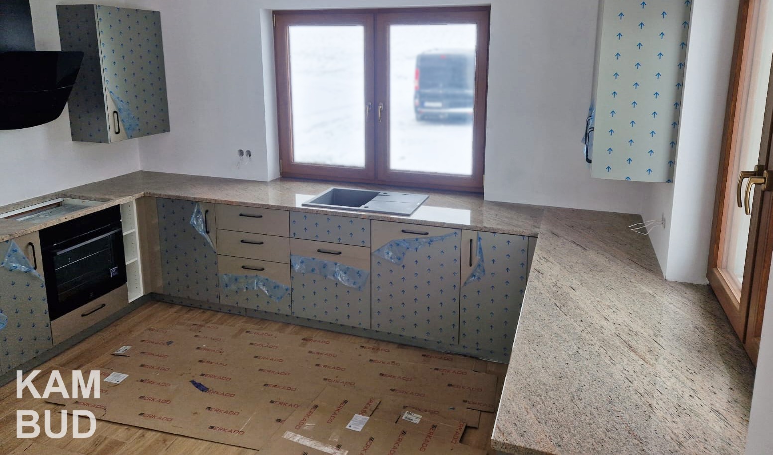 Montaż blatu kuchennego z jasnego kamienia, szafki zabezpieczone folią ochronną, widoczny zlew i okno z widokiem na zewnątrz, podłoga zabezpieczona kartonem.