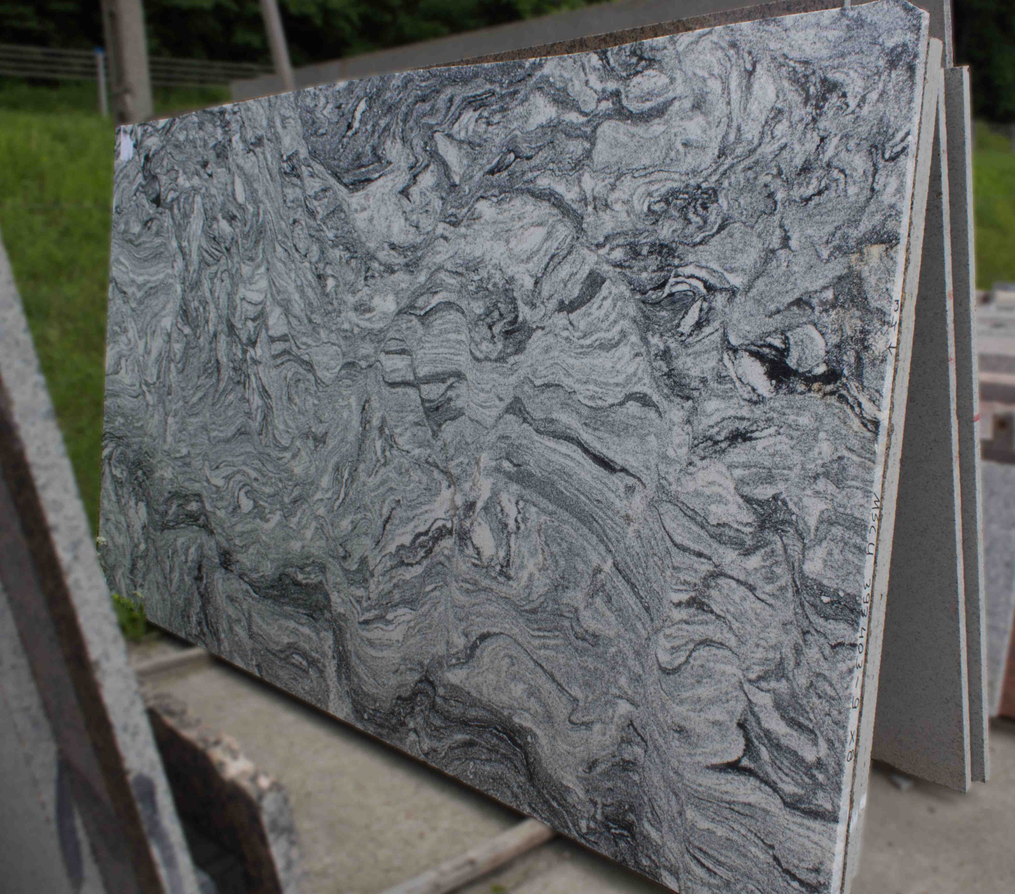 Płyta granitowa z wyraźnym, falistym wzorem w odcieniach szarości i bieli, oparta o inne płyty w magazynie kamieniarskim.