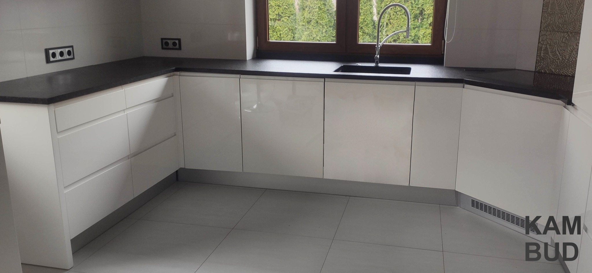 Nowoczesna kuchnia z białymi, lakierowanymi szafkami i ciemnym blatem, widok na zewnątrz przez okno nad zlewem, minimalistyczne gniazdka elektryczne na ścianie.