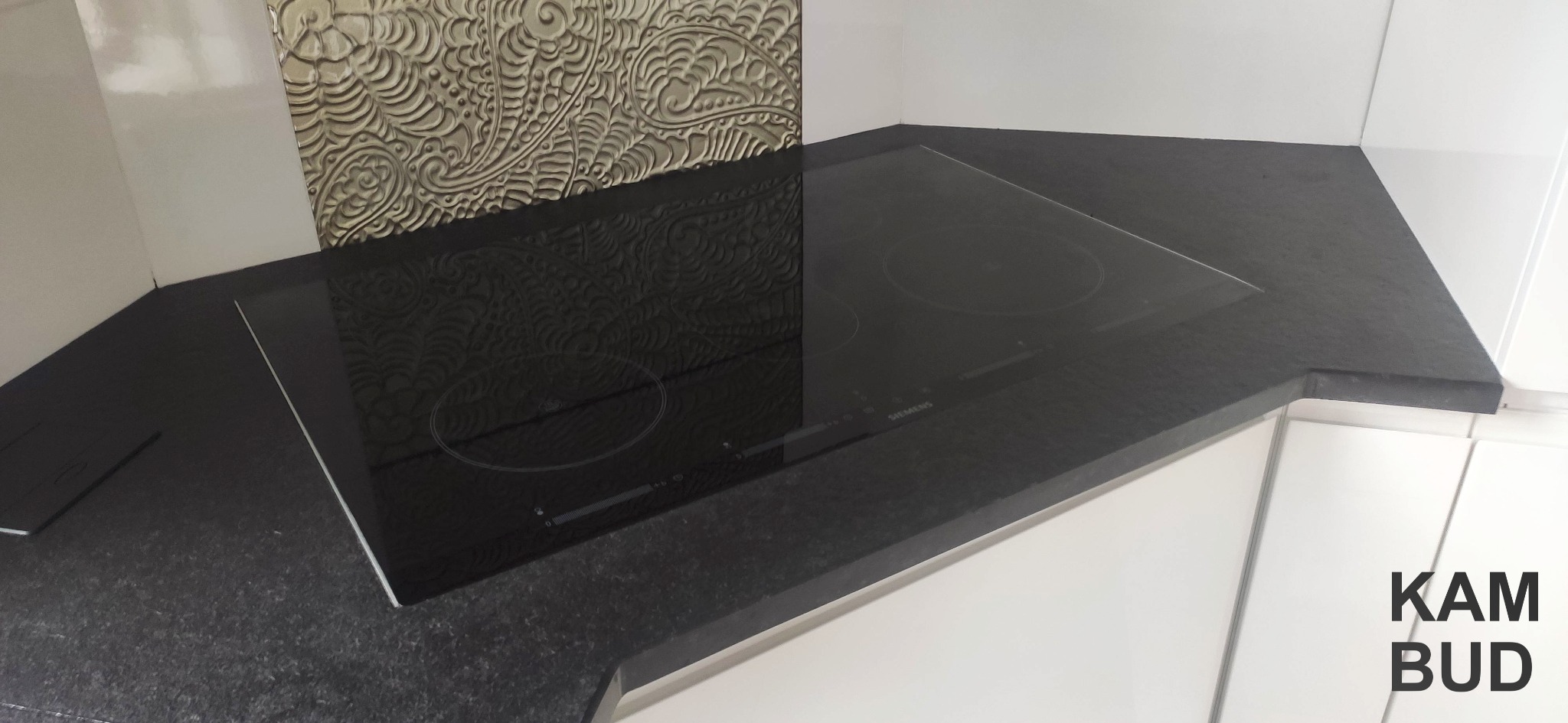 Czarny, granitowy blat kuchenny z zamontowaną płytą indukcyjną Siemens, na tle białych szafek i dekoracyjnej, beżowej płytki ściennej z wytłaczanym wzorem.