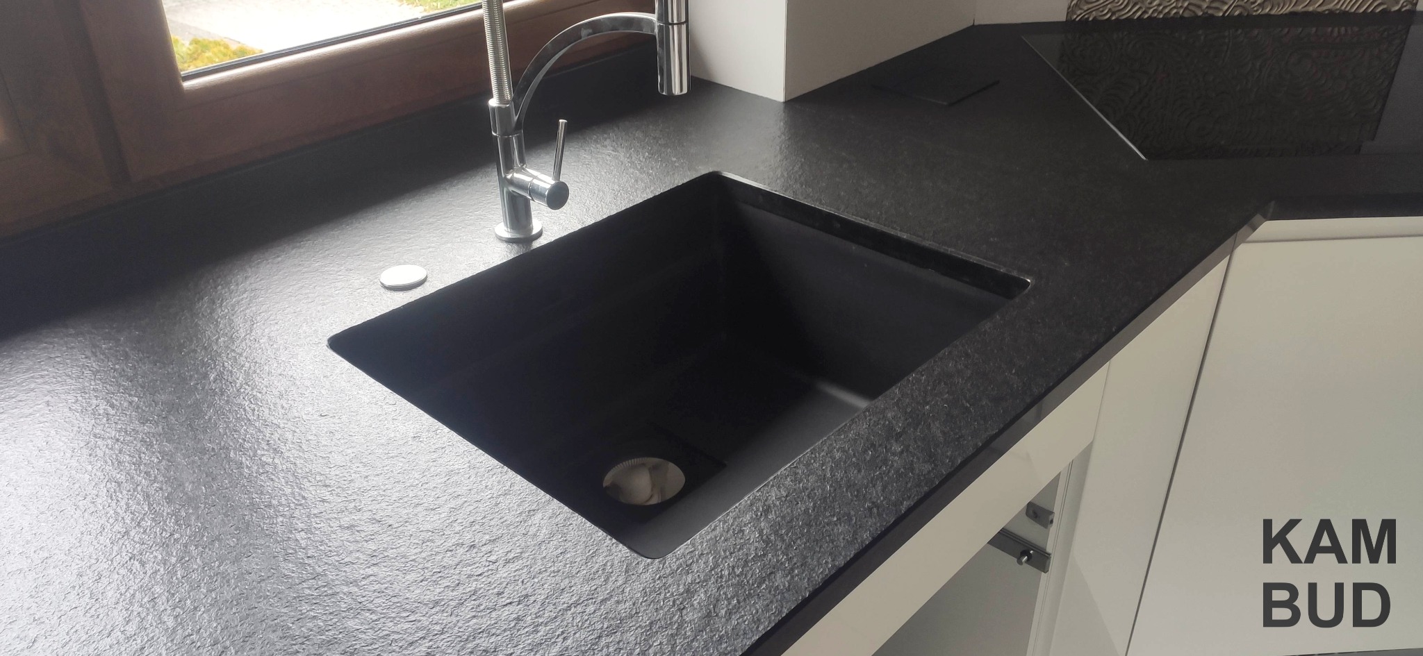 Czarny, teksturowany blat kuchenny z wbudowanym zlewem i nowoczesną baterią, oświetlony naturalnym światłem z okna, z widocznym logo firmy KAM BUD na białej szafce.