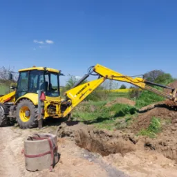 Żółta koparko-ładowarka Fermec podczas prac ziemnych, z wykopanym rowem i stosem ziemi, na tle błękitnego nieba i zielonych pól.