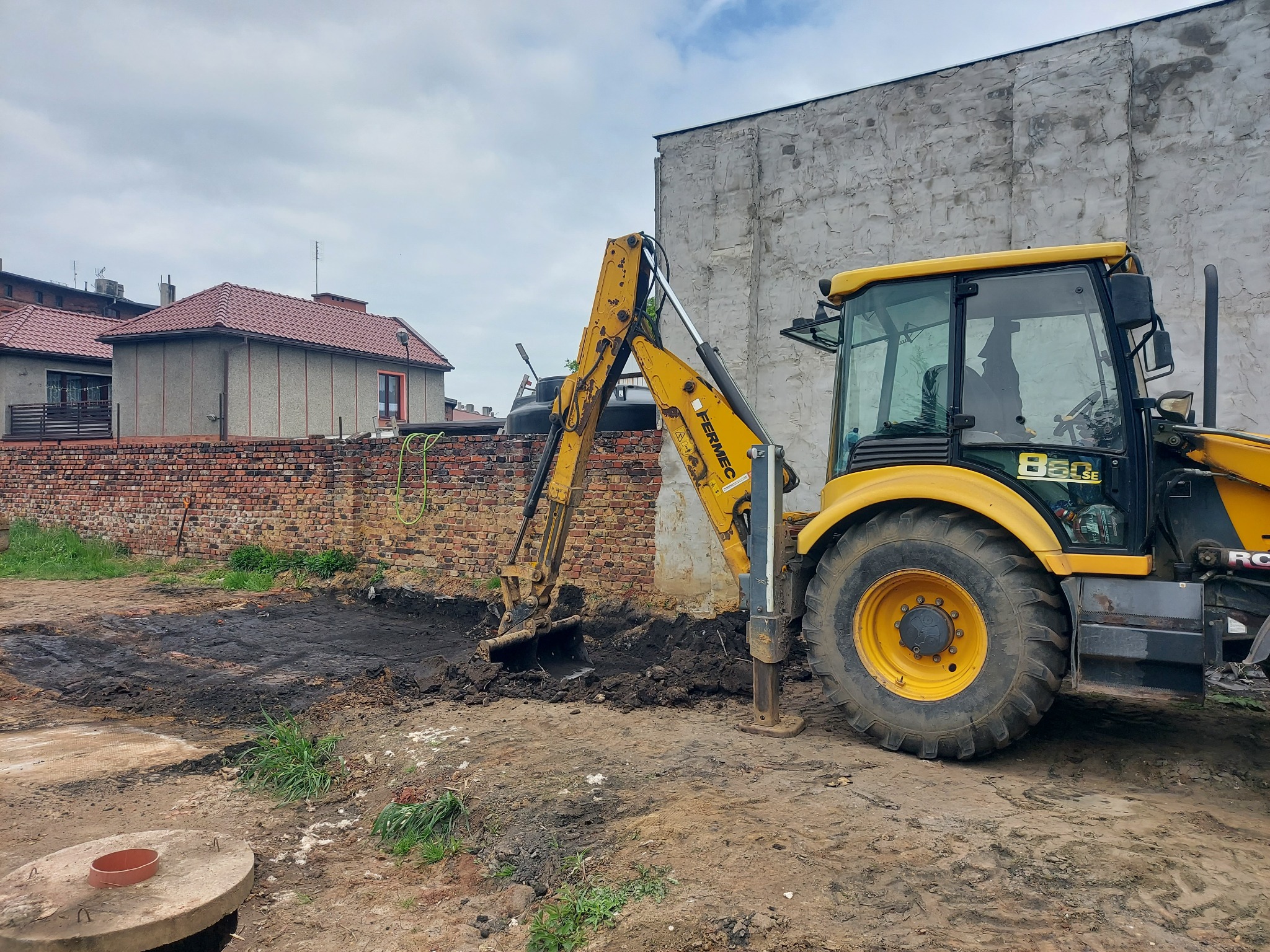 Żółta koparko-ładowarka Fermec 860SE w trakcie prac ziemnych, widoczny wykop w gruncie obok ceglanego muru i betonowego budynku, pochmurne niebo.