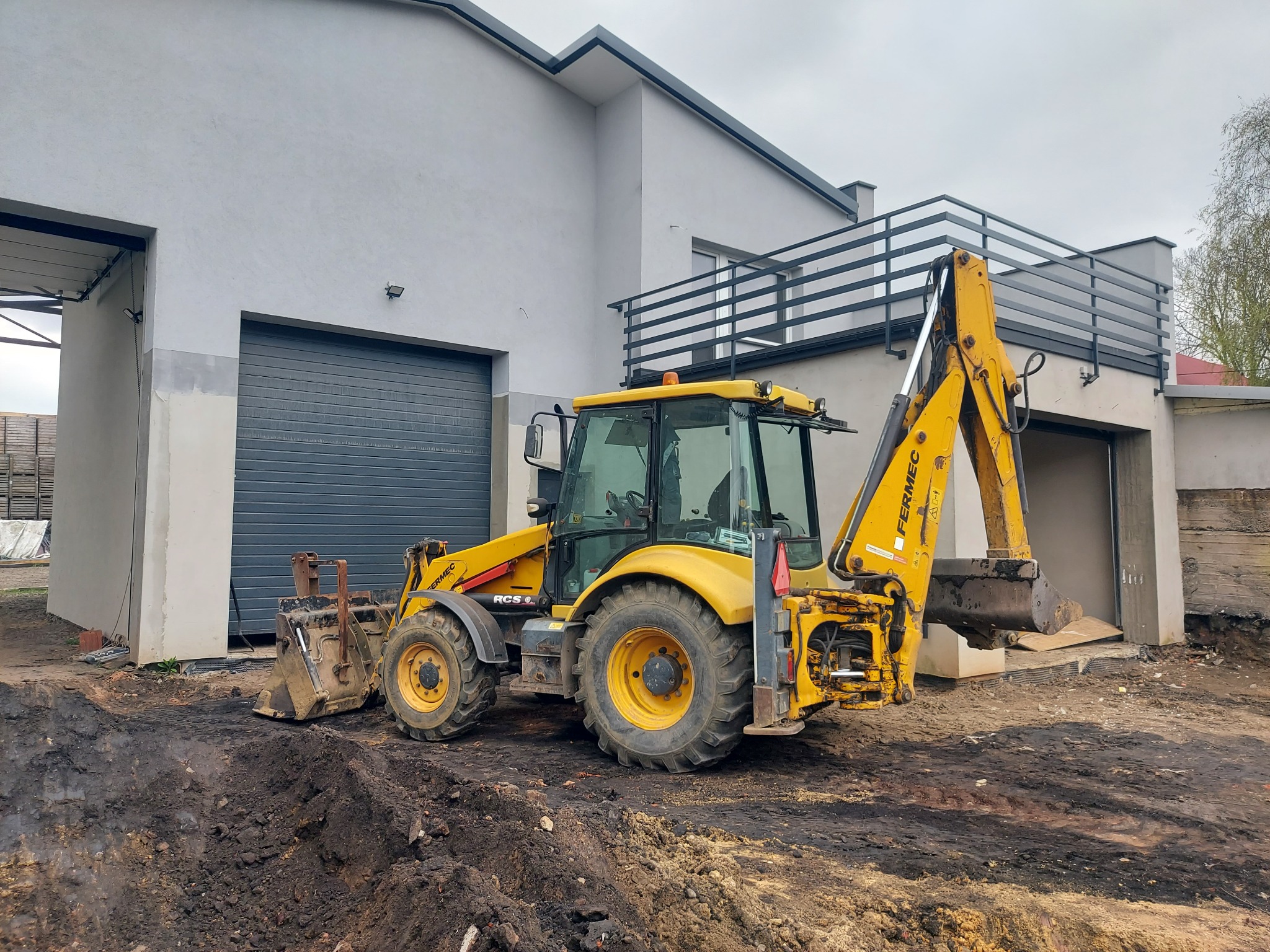 Żółta koparko-ładowarka Fermec RCS w trakcie prac ziemnych na placu budowy, z widocznym wykopem i nowym budynkiem w tle.