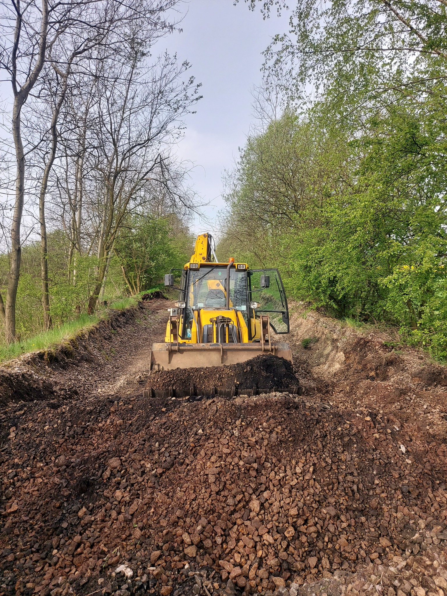 Żółta koparko-ładowarka JCB na wykopie w terenie zalesionym, widoczne świeżo usunięte warstwy ziemi i kamieni.