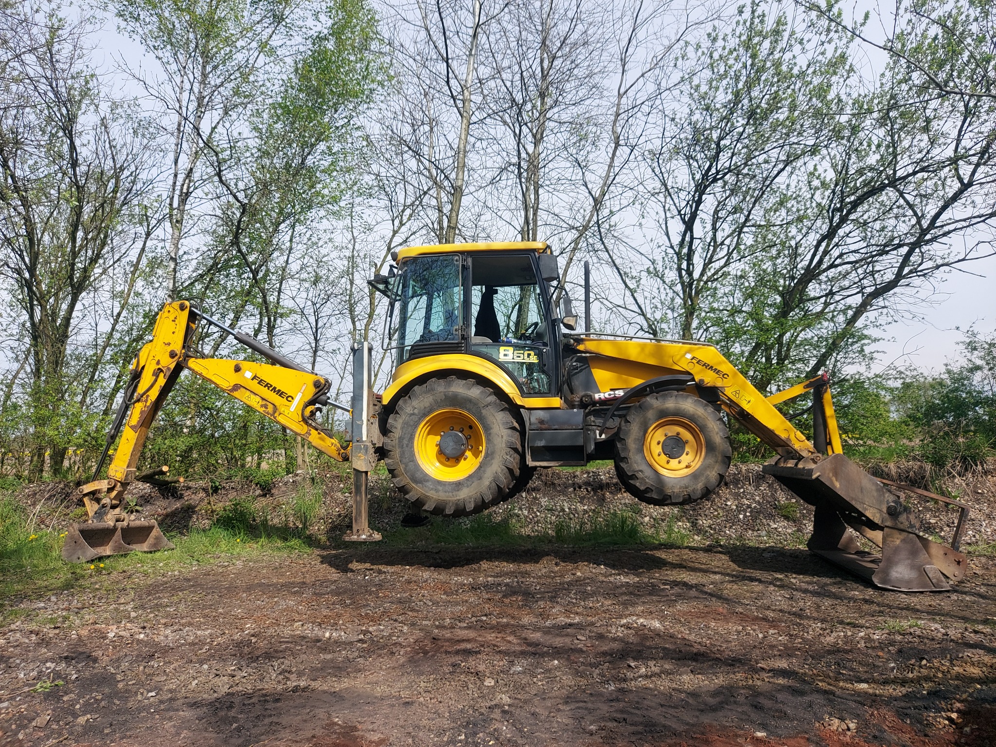 Żółta koparko-ładowarka marki Fermec, model 860, zawieszona w powietrzu podczas stabilizacji na podporach, na tle drzew bez liści i ziemi.