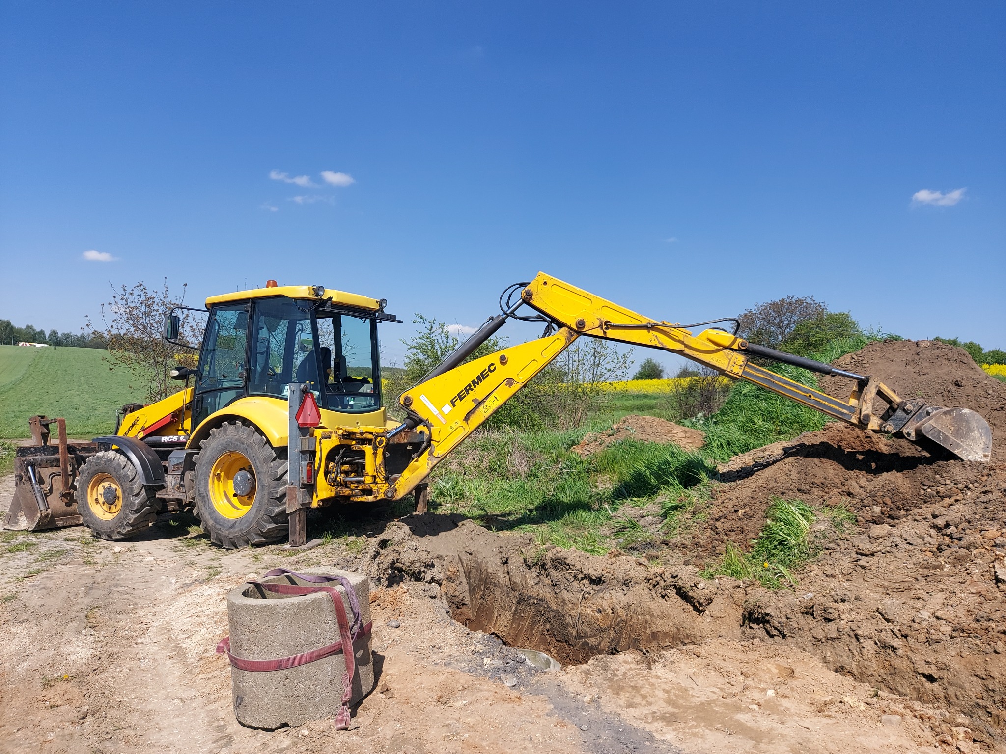 Żółta koparko-ładowarka Fermec podczas prac ziemnych, z wykopanym rowem i stosem ziemi, na tle błękitnego nieba i zielonych pól.
