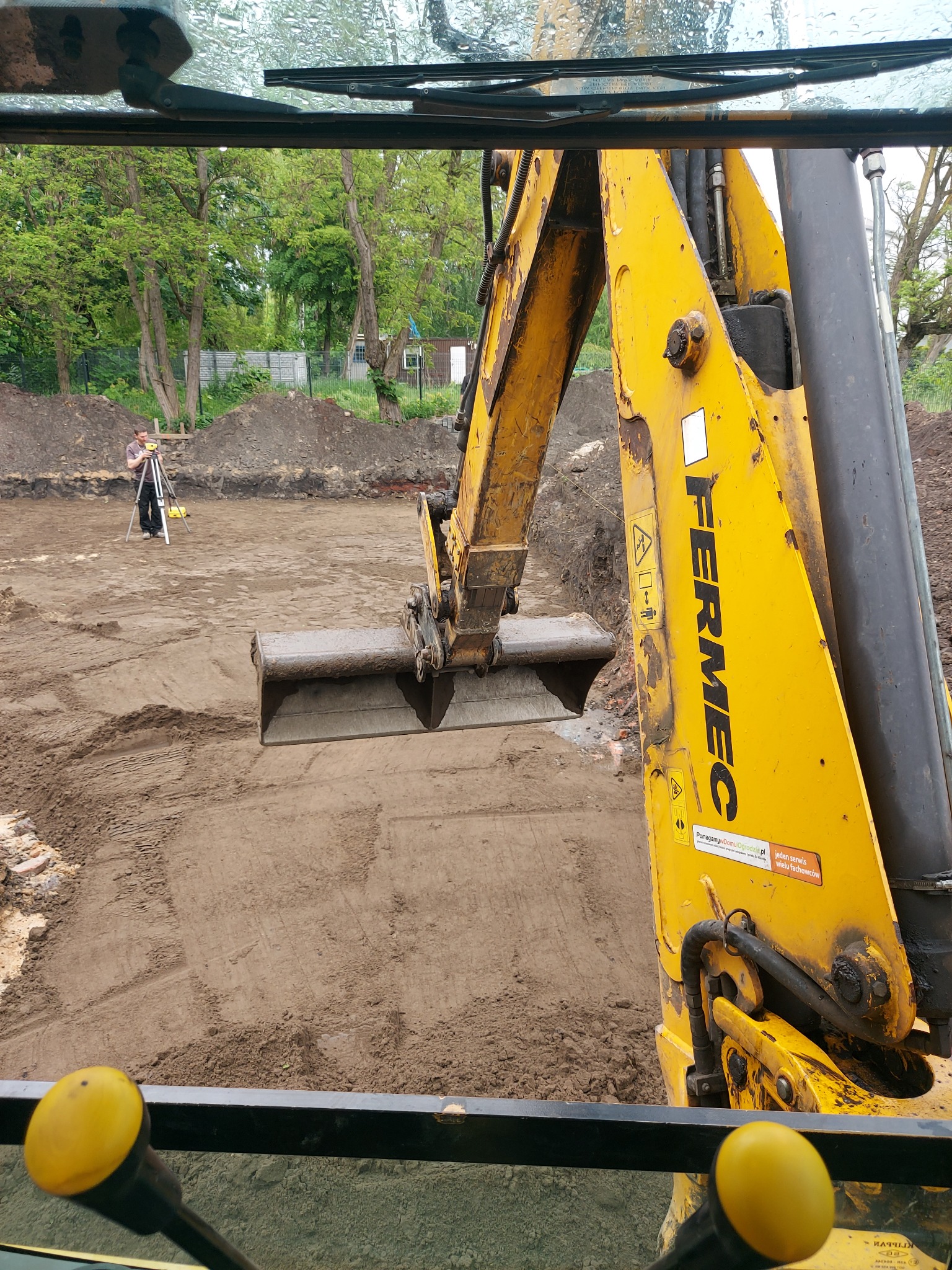 Widok z kabiny koparki na wykop pod fundamenty, w tle geodeta z niwelatorem, żółty wysięgnik koparki Fermec