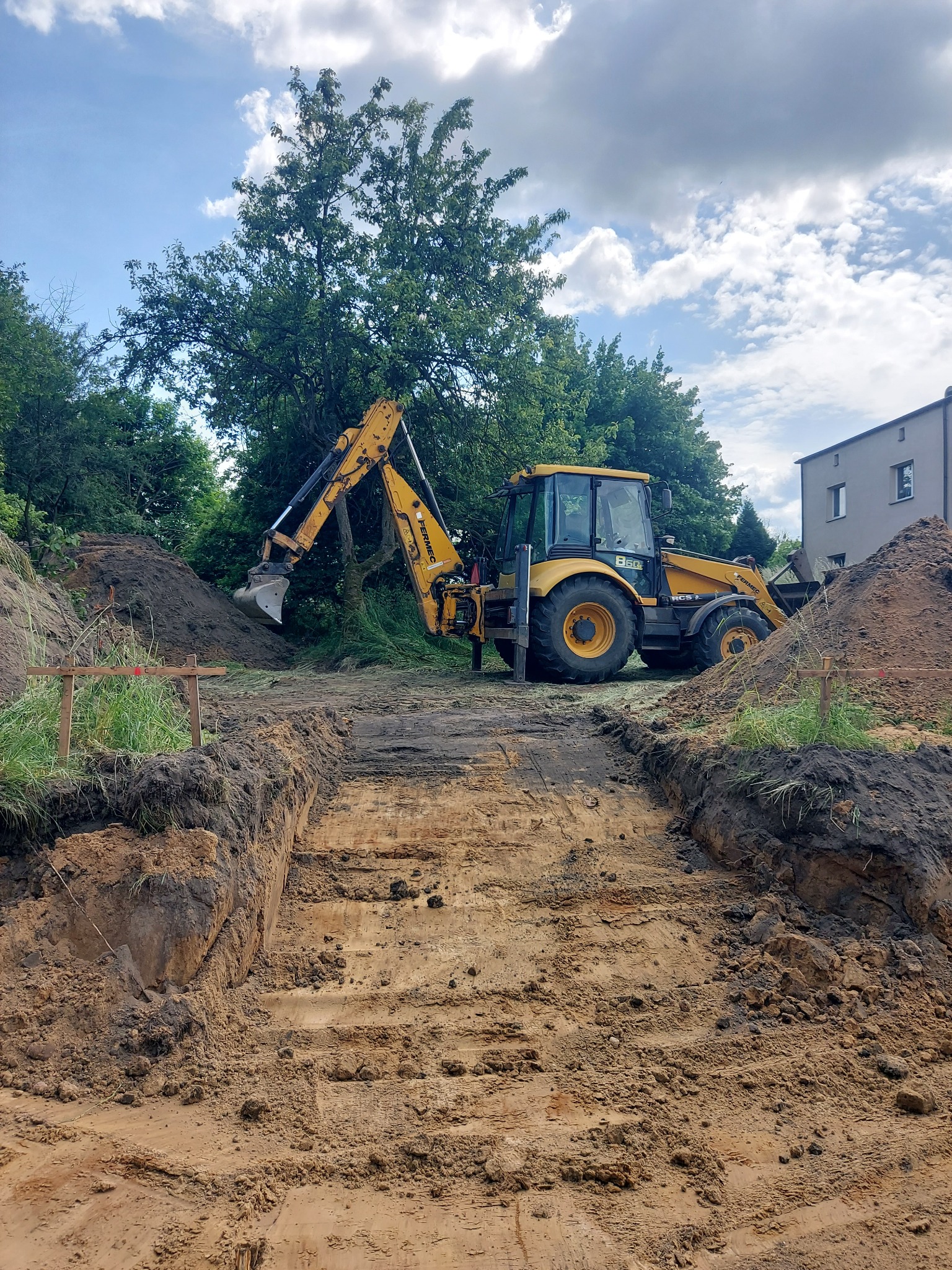 Żółta koparko-ładowarka Fermec 860CS przy wykopie w ziemi, pryzmy ziemi po bokach, w tle budynek i drzewa pod błękitnym niebem z chmurami.