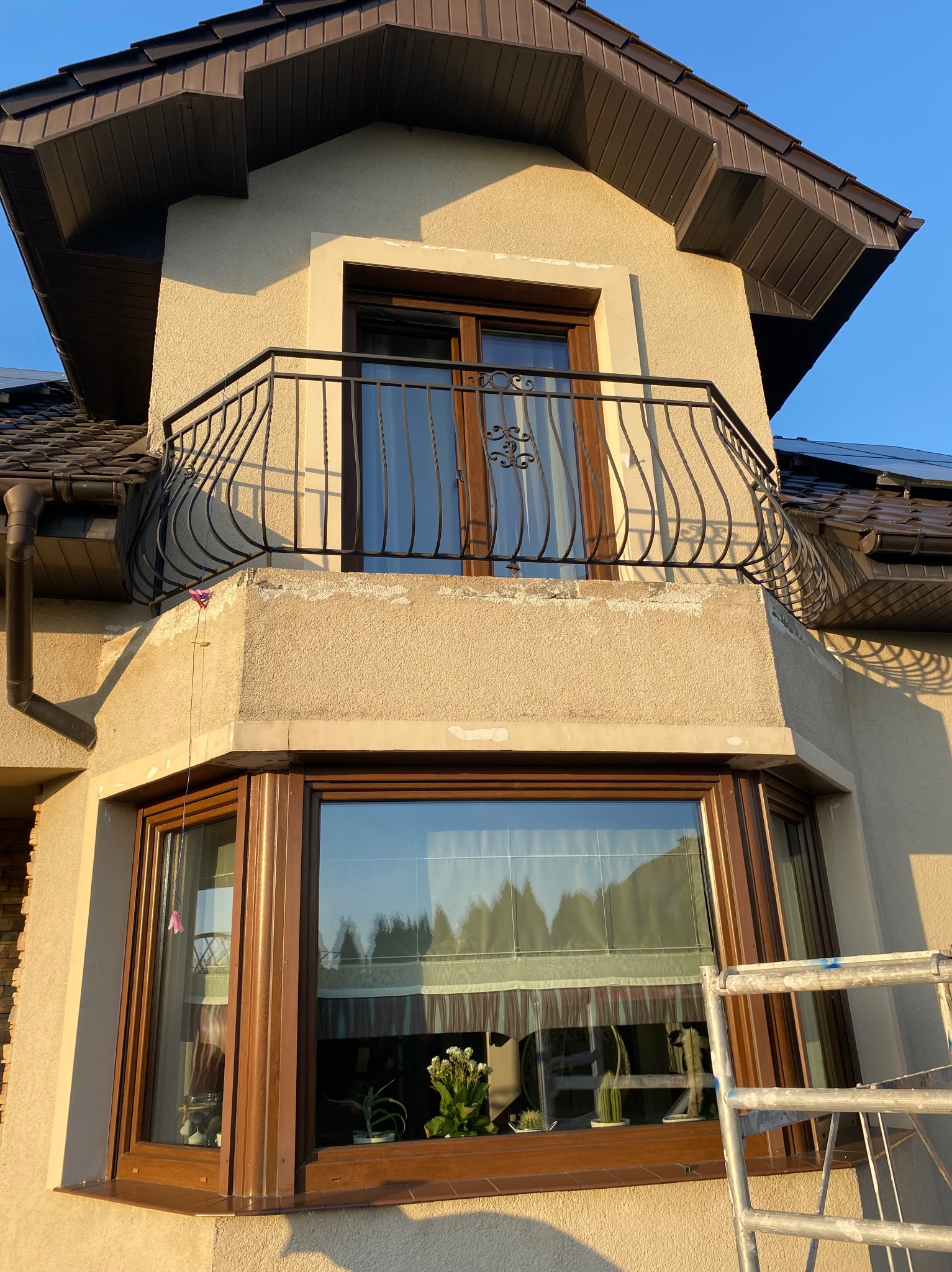 Fragment fasady domu z balkonem i oknem wykuszowym, widoczne ślady po pracach wykończeniowych, rusztowanie przy oknie, elewacja w odcieniach beżu, brązowe okna i dachówka.