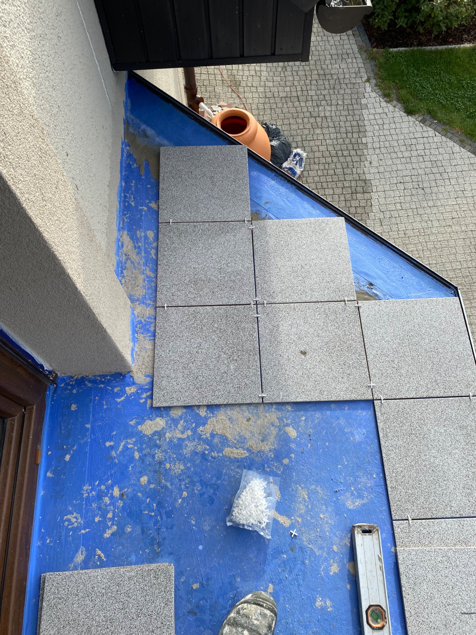 Montaż szarych płytek na balkonie z użyciem krzyżyków dystansowych i poziomicy, widoczna niebieska hydroizolacja i fragment buta roboczego.