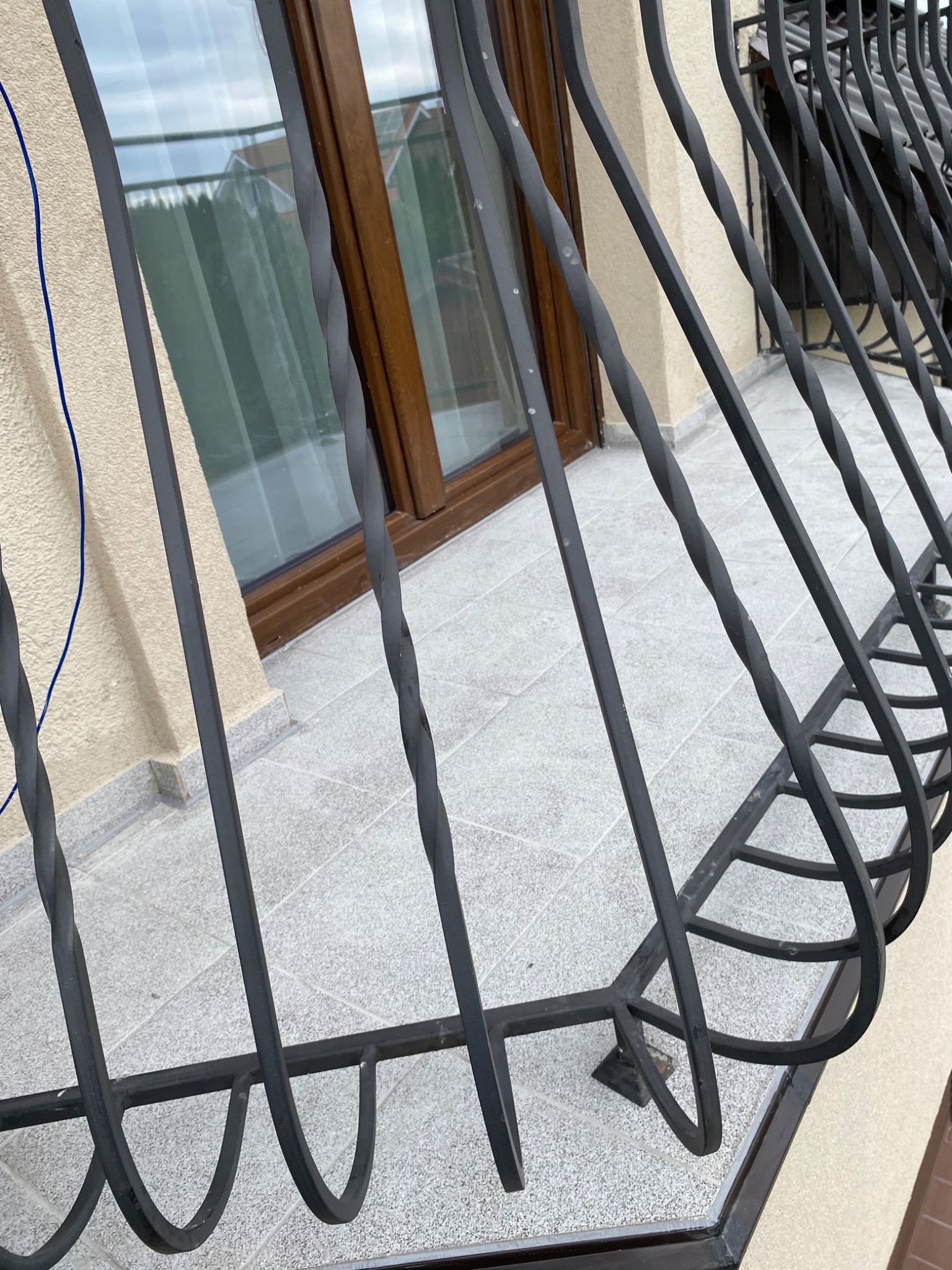 Kute, czarne balustrady balkonowe z ozdobnymi skręceniami na tle beżowej elewacji i okna z brązową ramą.