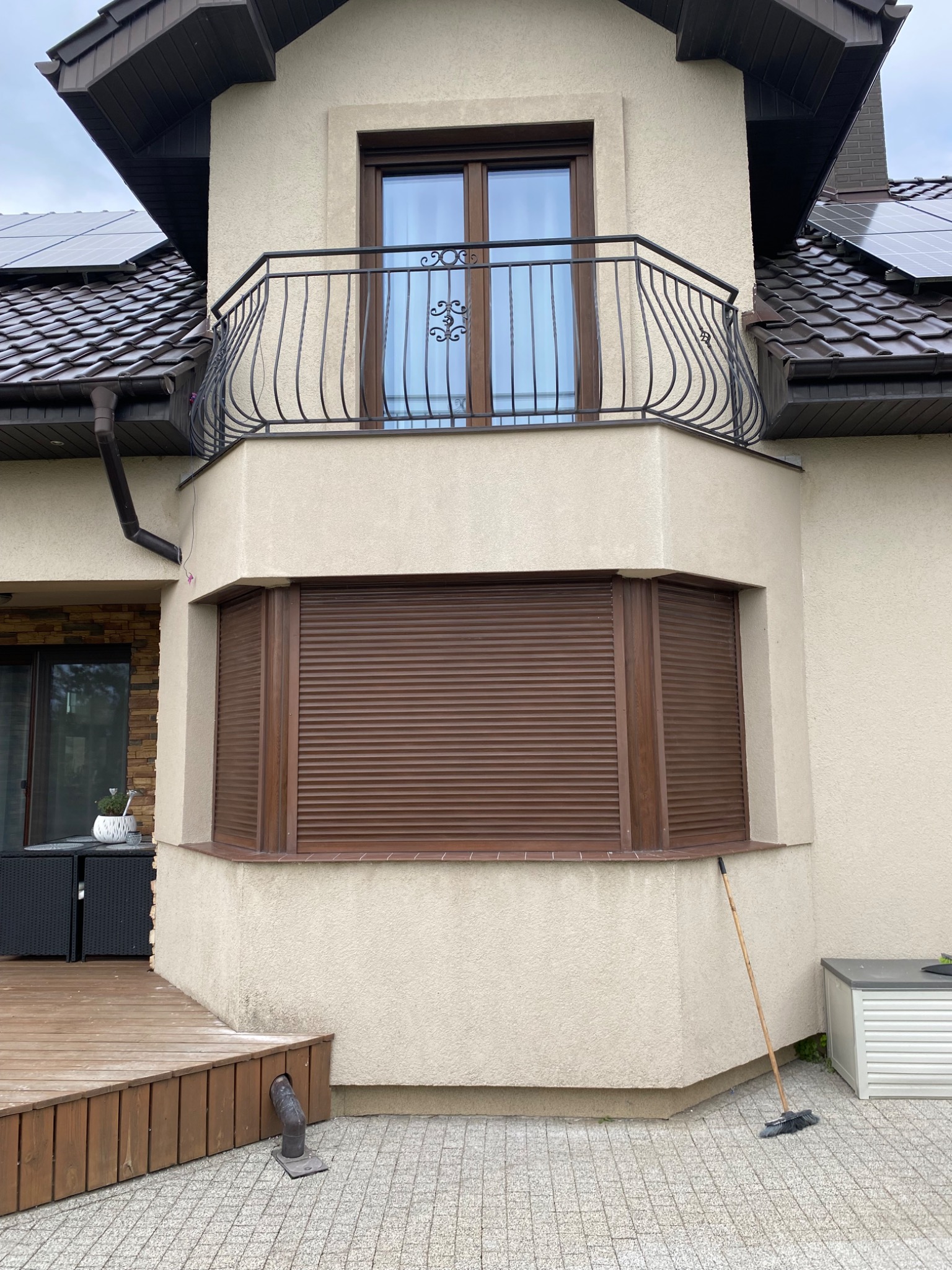 Fragment elewacji domu z balkonem, brązowymi roletami zewnętrznymi na oknach i panelami słonecznymi na dachu, widok z poziomu chodnika.