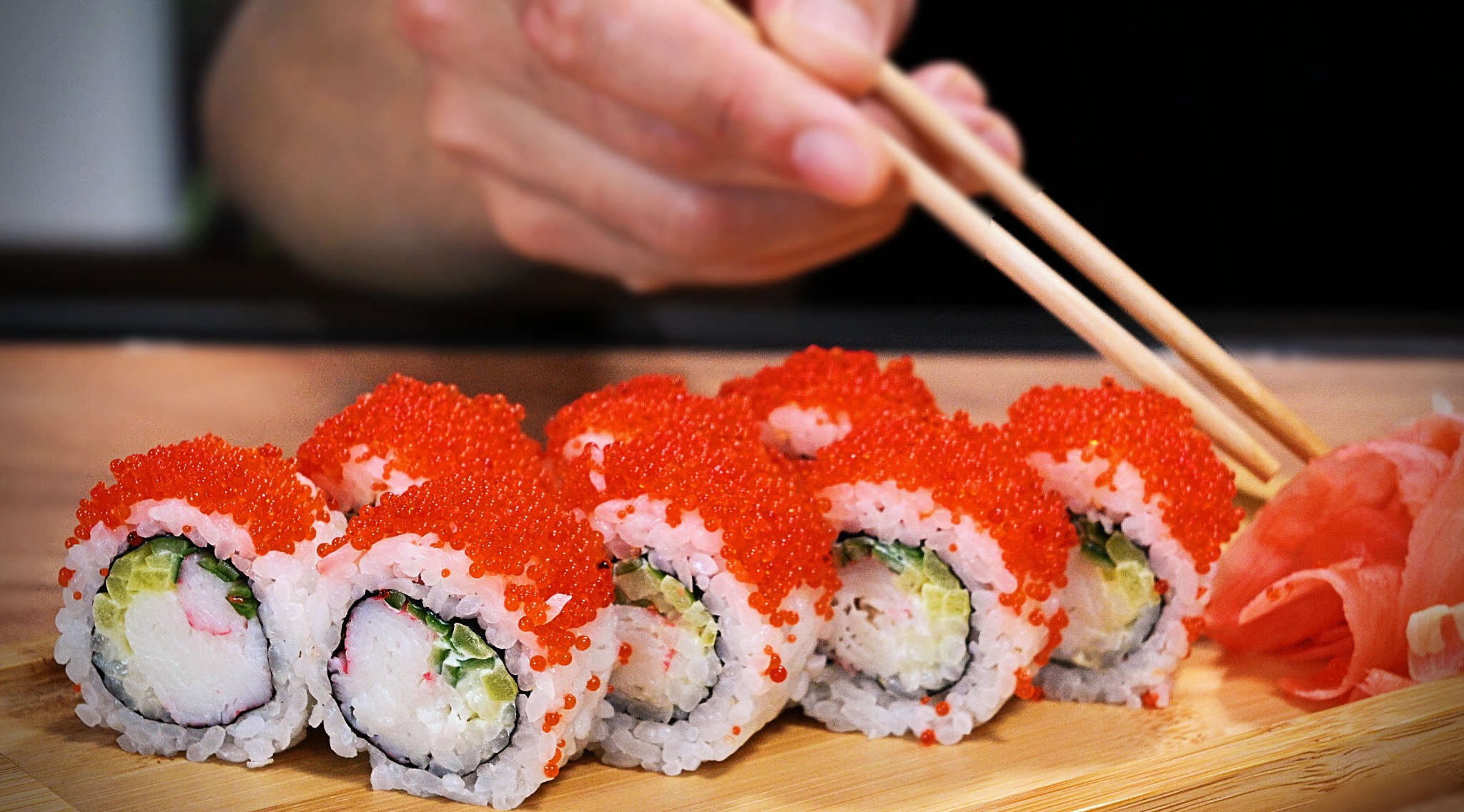 Szereg rolek sushi California z czerwonym kawiorem na drewnianej desce, zbliżające się pałeczki i marynowany imbir w tle.