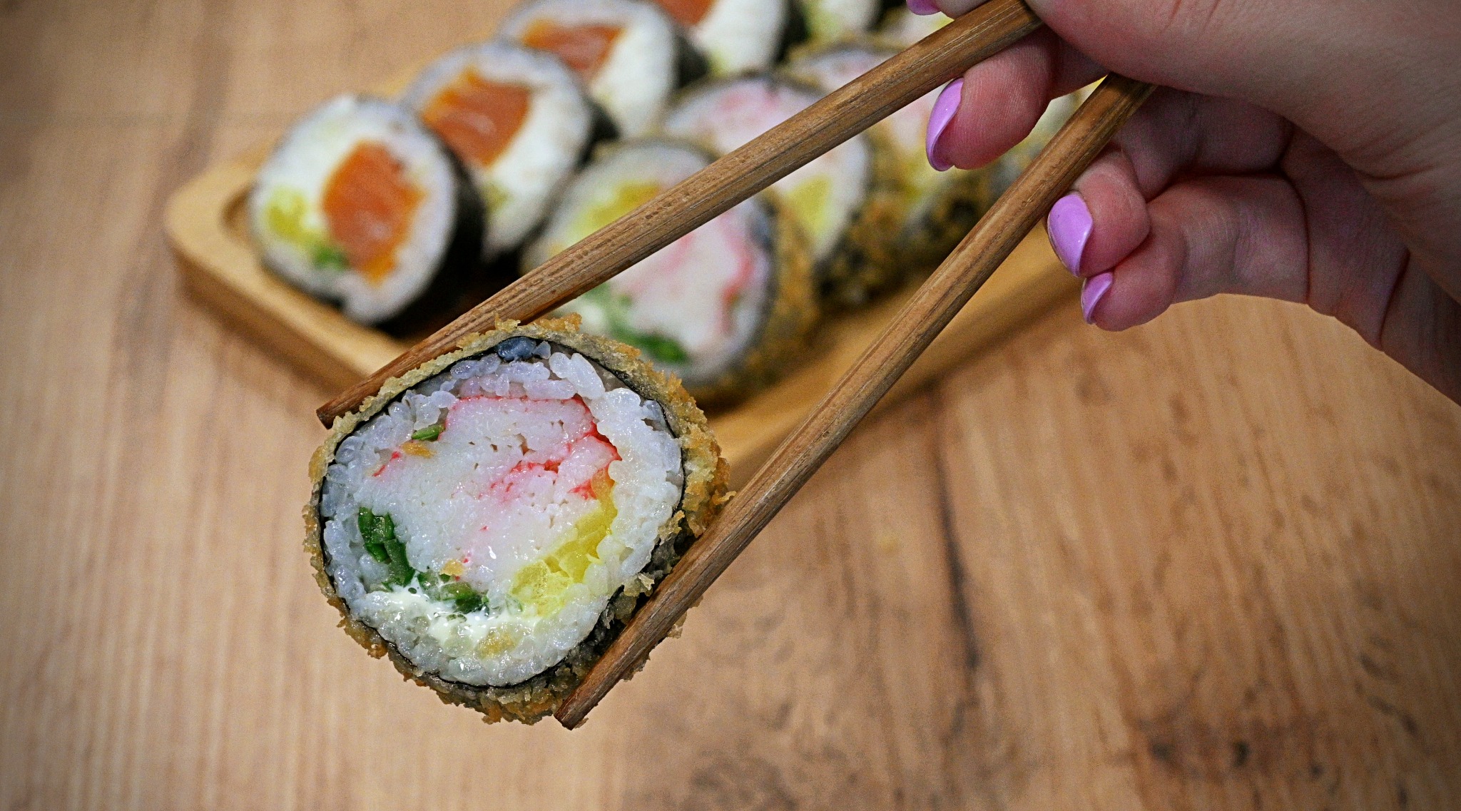 Smażona rolka sushi z krabem, ogórkiem i serkiem, podnoszona pałeczkami, na drewnianej desce z innymi rolkami w tle, dłoń z fioletowym manicure.